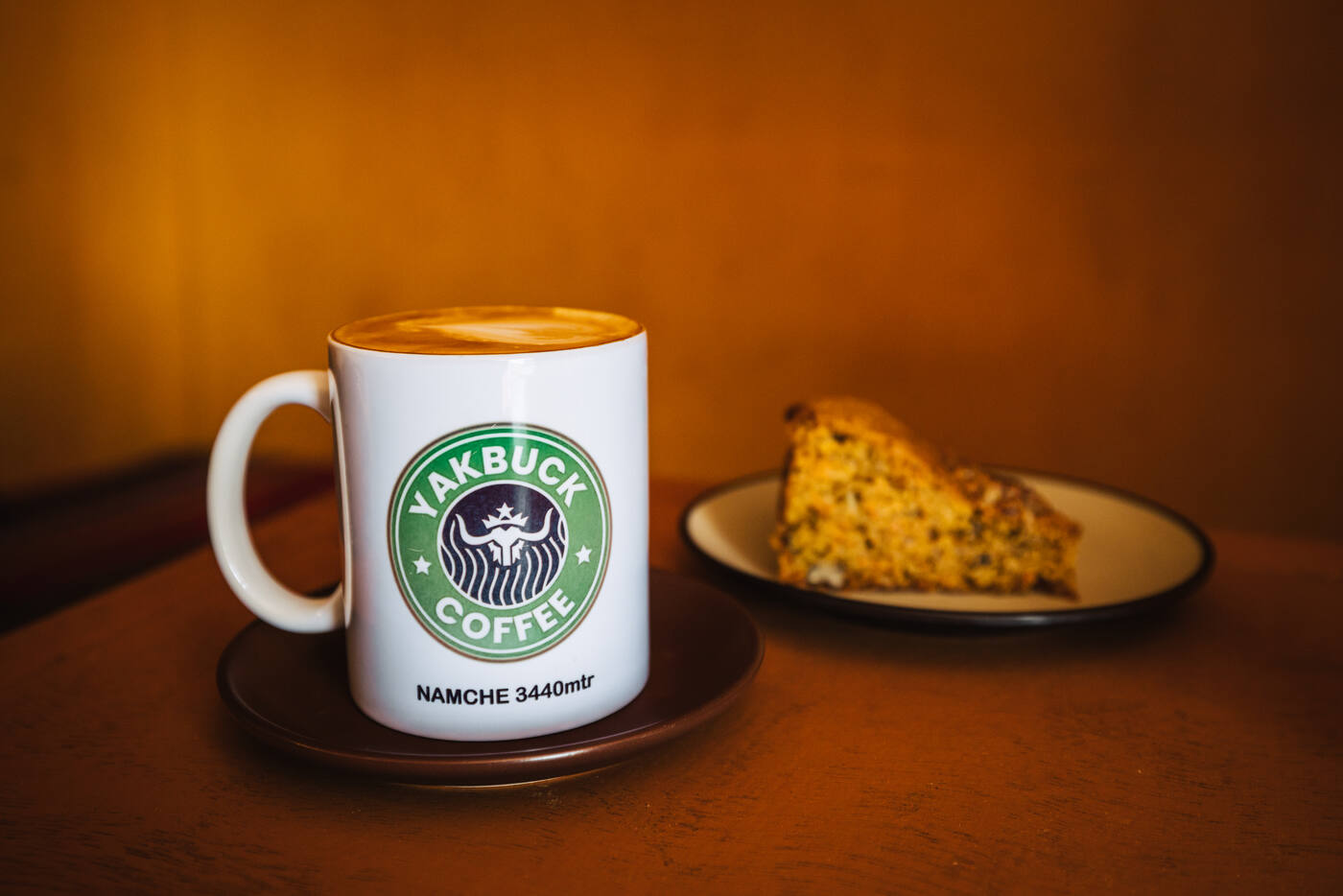 A Yakbuck Coffee latte on a saucer beside a slice of carrot cake — Namche 3,440m printed on the mug