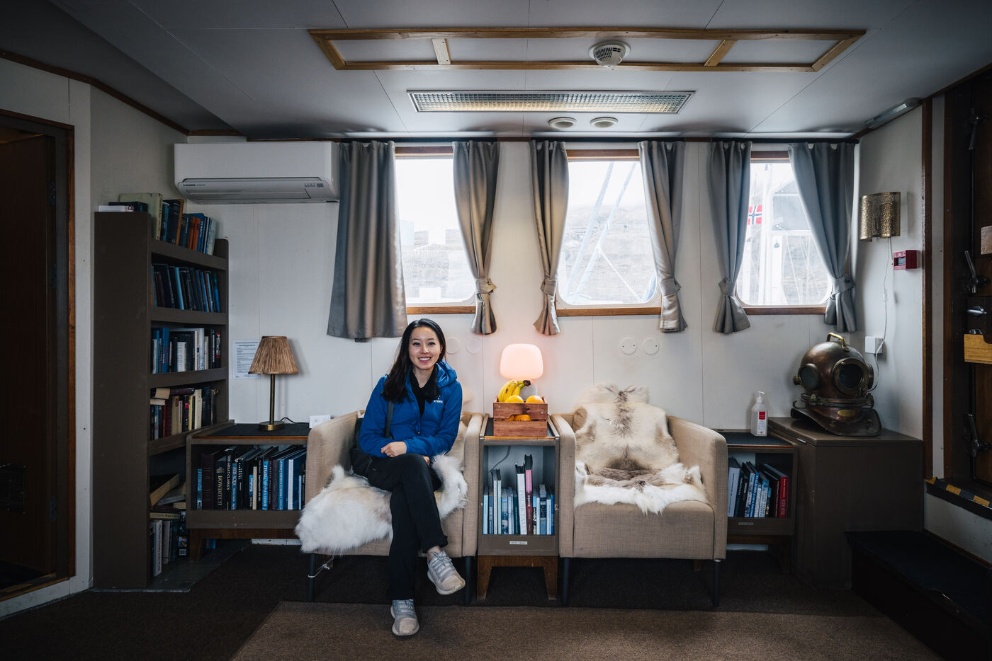 Woman in a blue jacket seated in the Kinfish lounge between bookshelves and reindeer-hide armchairs