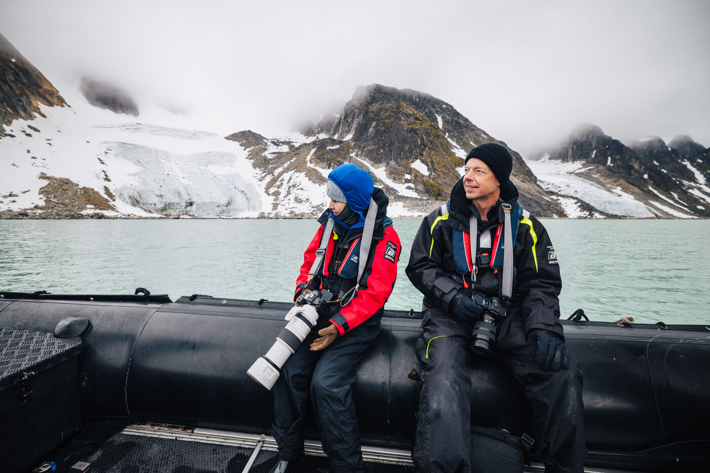 Two passengers with cameras seated on the zodiac pontoon, glacier and peaks behind