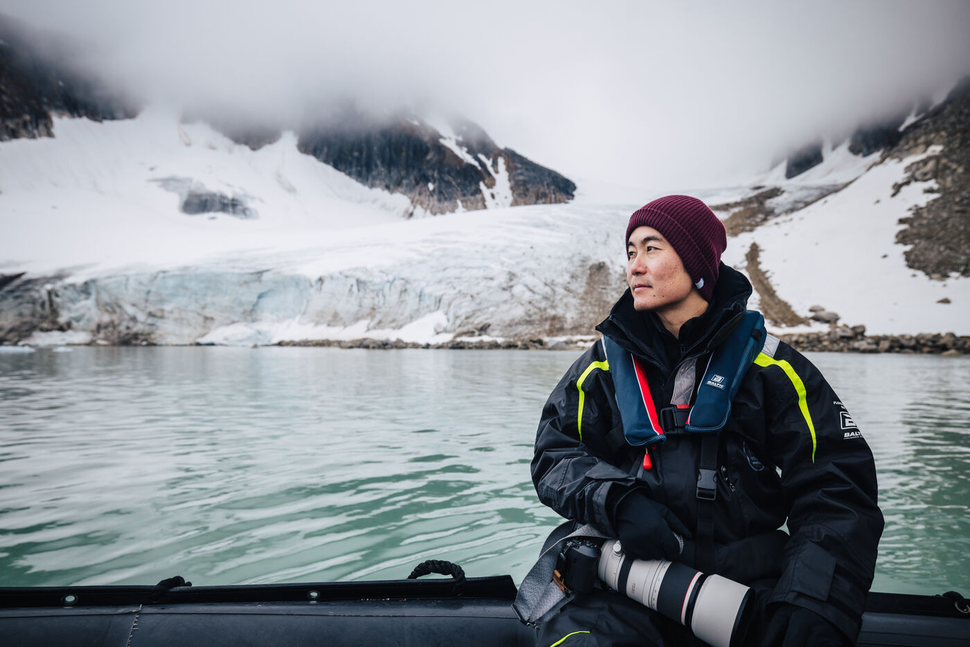 Man in a maroon beanie seated in the zodiac with a telephoto lens, glacier front behind