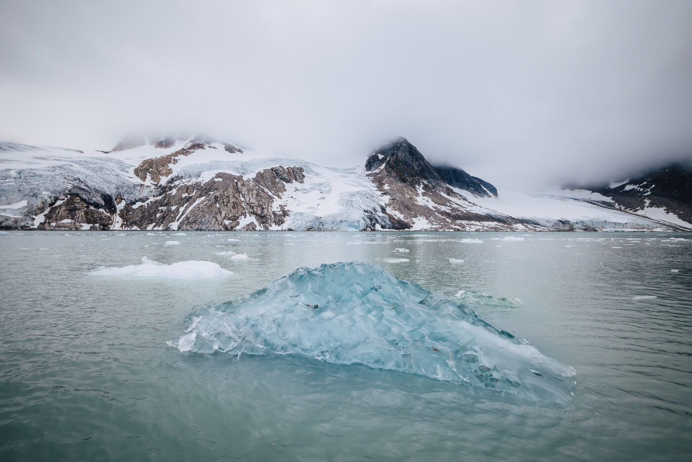 Clear iceberg floating in front of a glacier and cloud-capped peaks