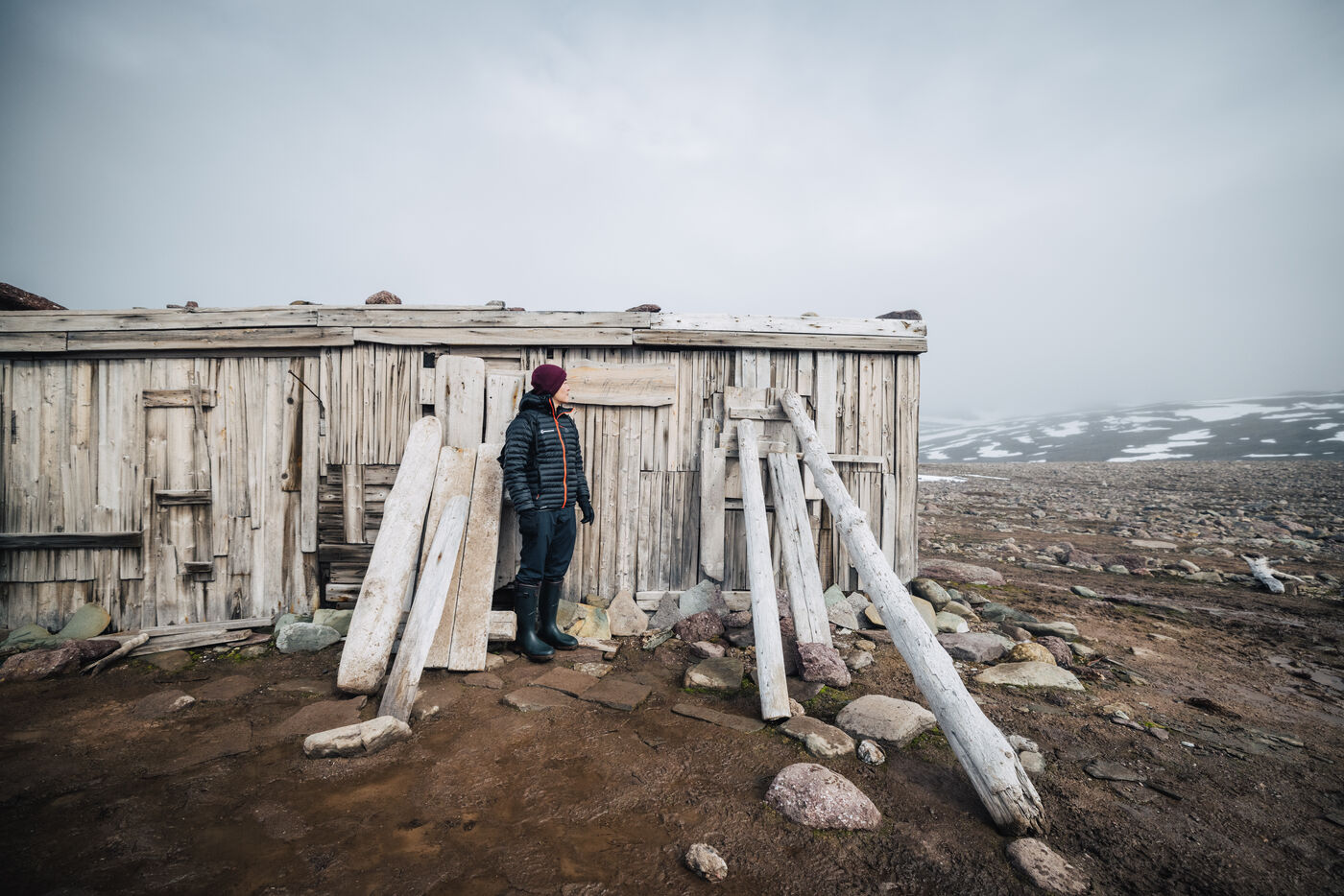Person standing near a dilapidated wooden structure with snowy mountains