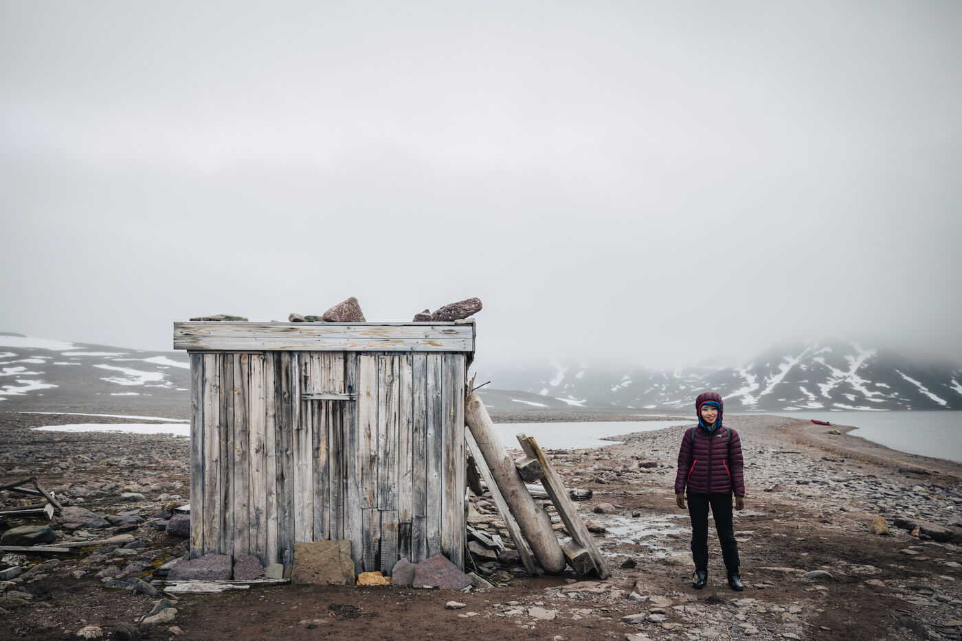 Person in a dark puffer standing beside the weathered trapper's hut
