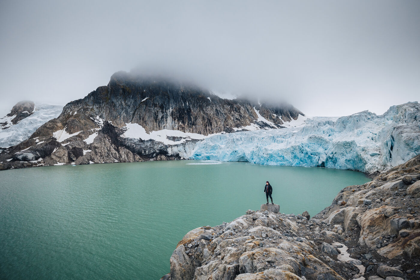 Lone figure on a rock outcrop above a turquoise glacial lagoon, glacier front beyond