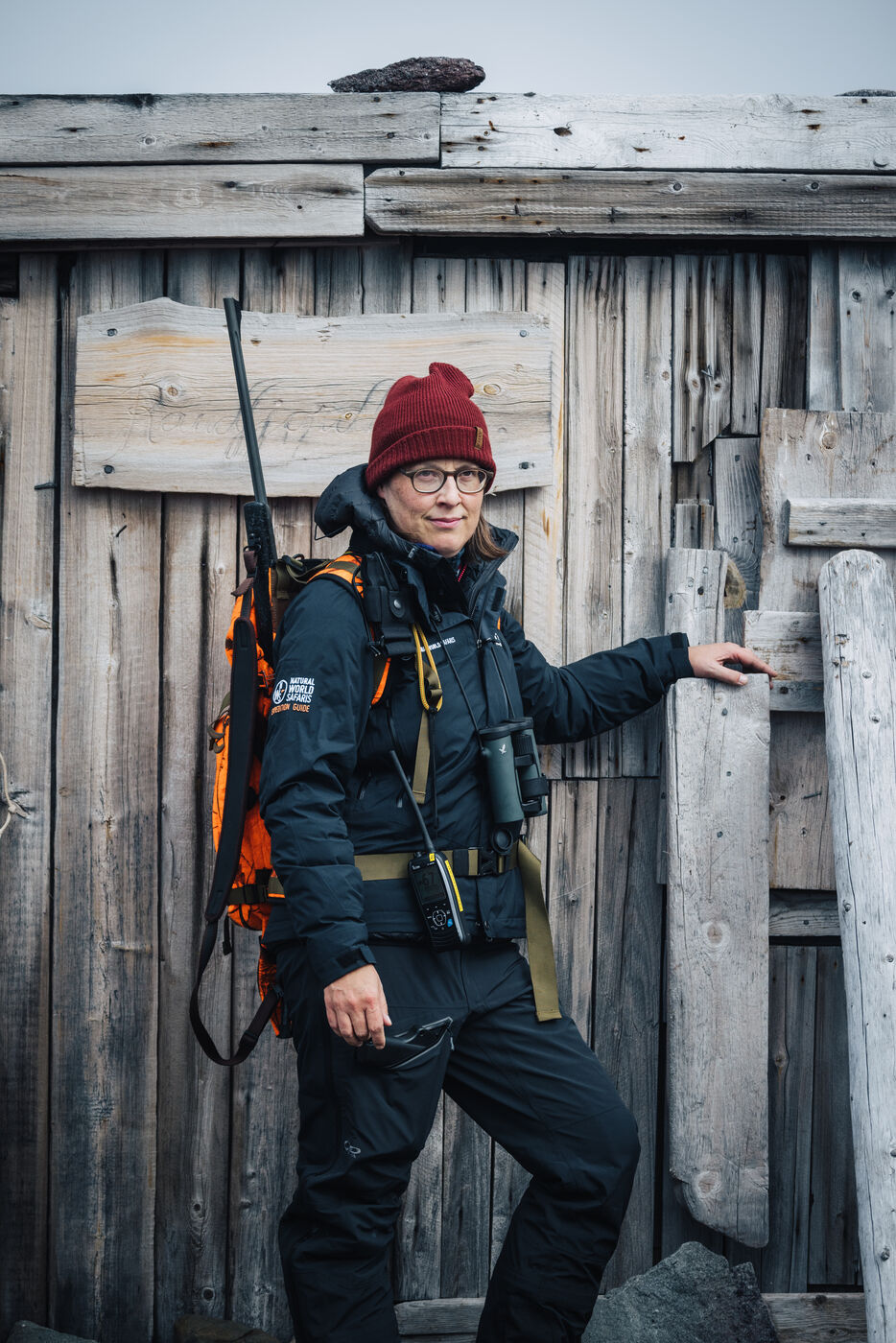 Guide with a rifle slung over his shoulder leaning against the trapper's hut