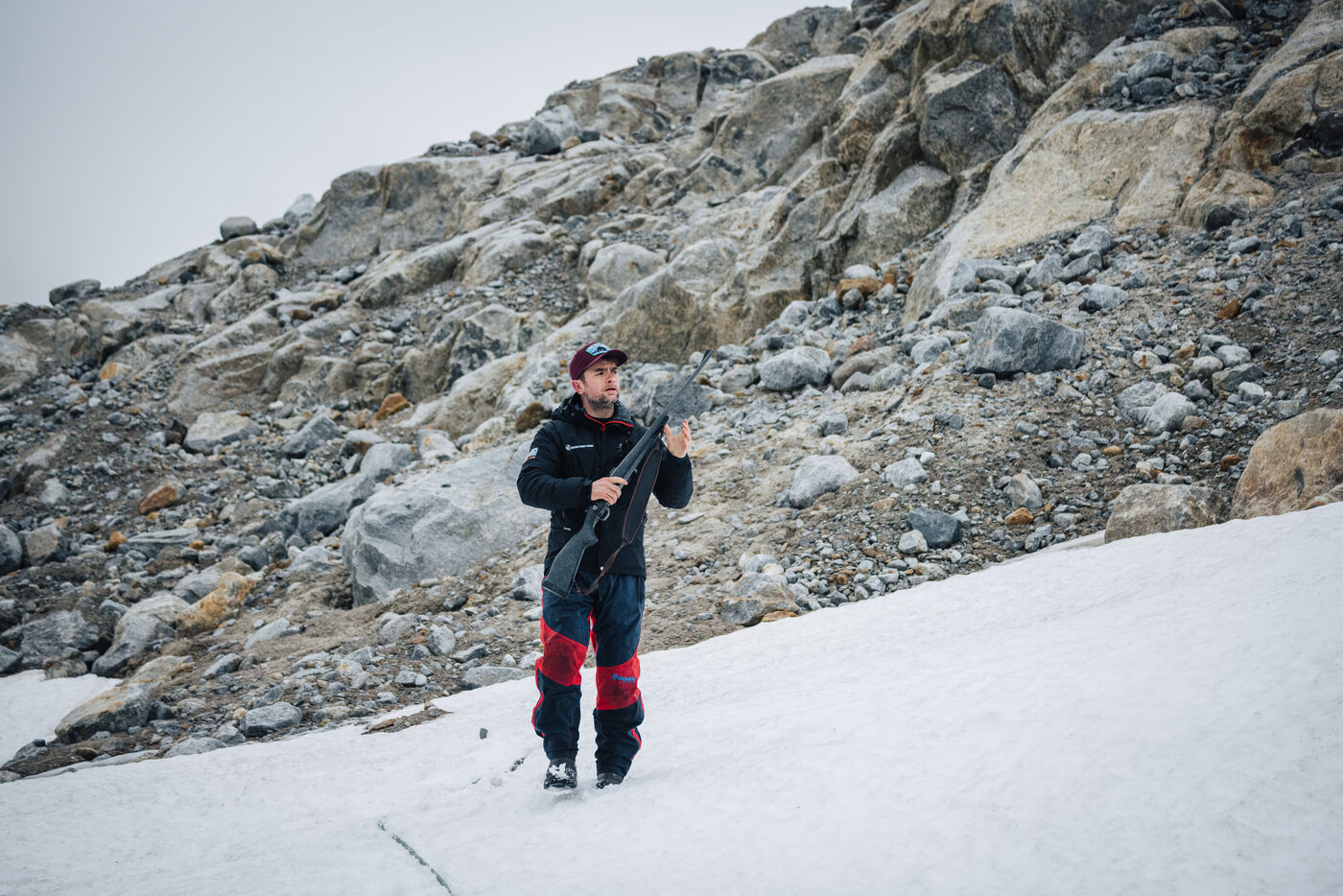 Guide carrying a rifle across a snow patch below a scree slope
