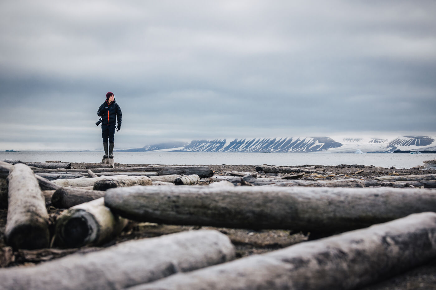 Person standing among driftwood logs on the beach, snow-striped mountains across the water