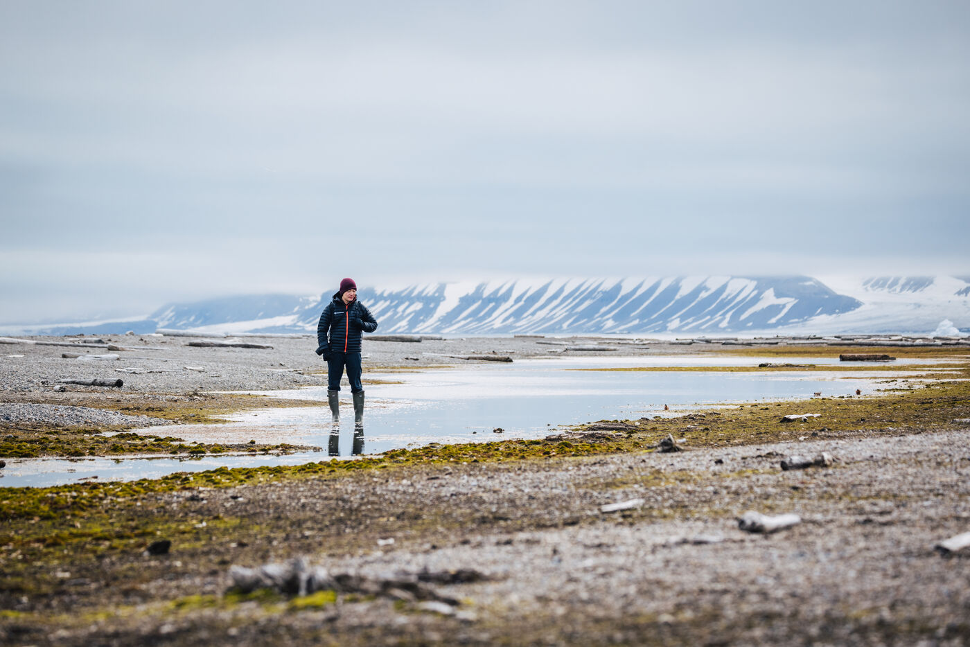 Person standing on wet tundra with snow-streaked mountains across the water