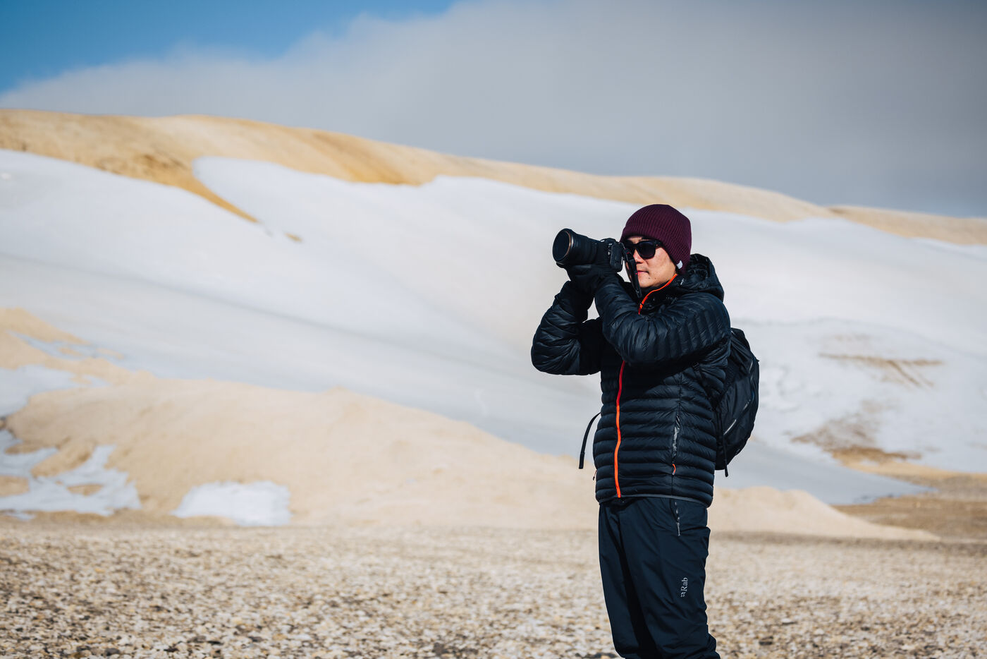 Person in a maroon beanie photographing with a telephoto lens, snow-covered hills behind
