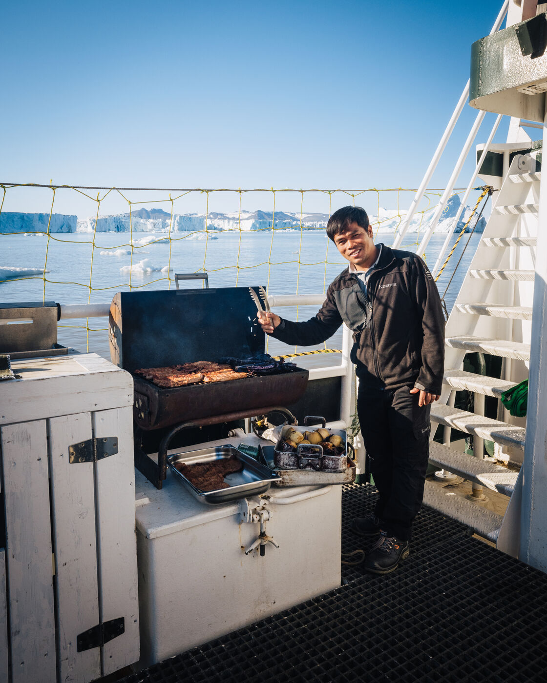 Crew member grilling on the Kinfish's aft deck with icebergs beyond