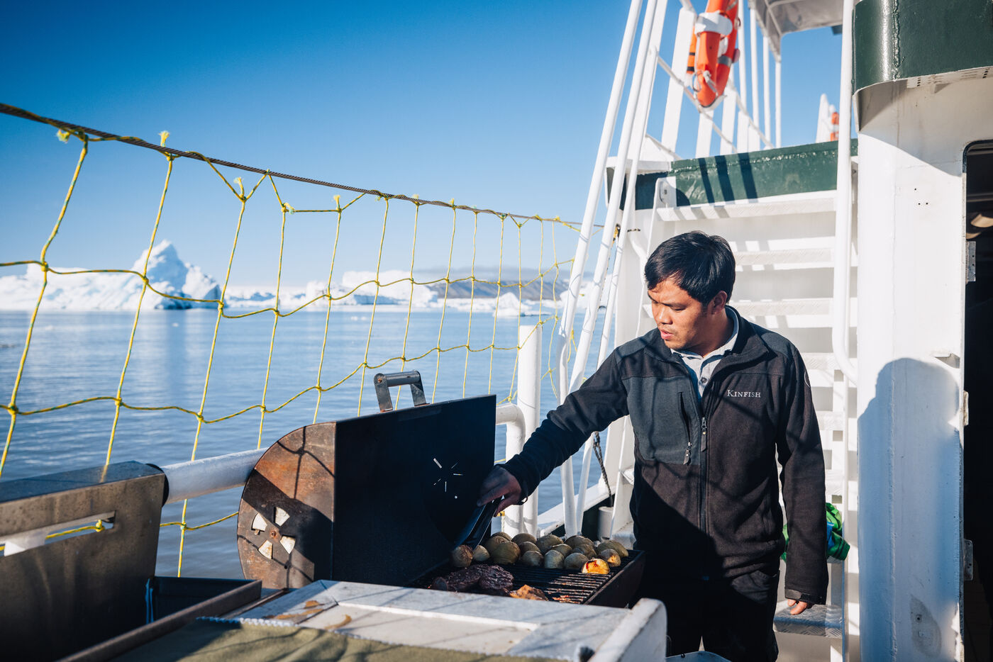 Crew member tending the deck barbecue, an iceberg framed behind him
