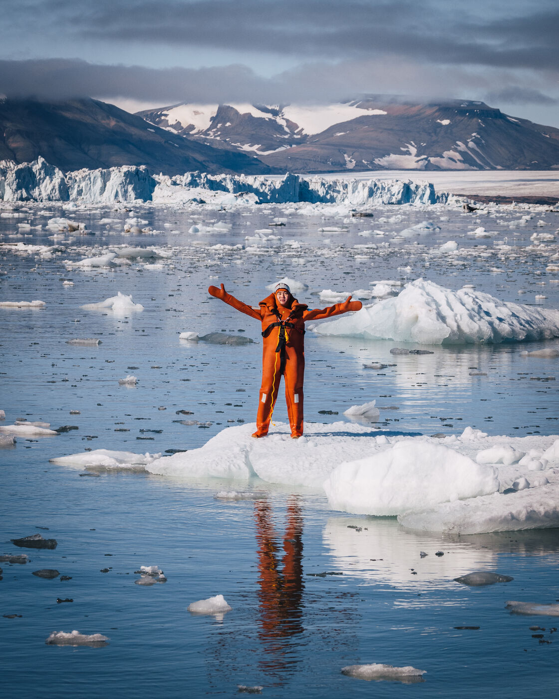 Person in an orange survival suit standing arms outstretched on an ice floe, glacier front behind