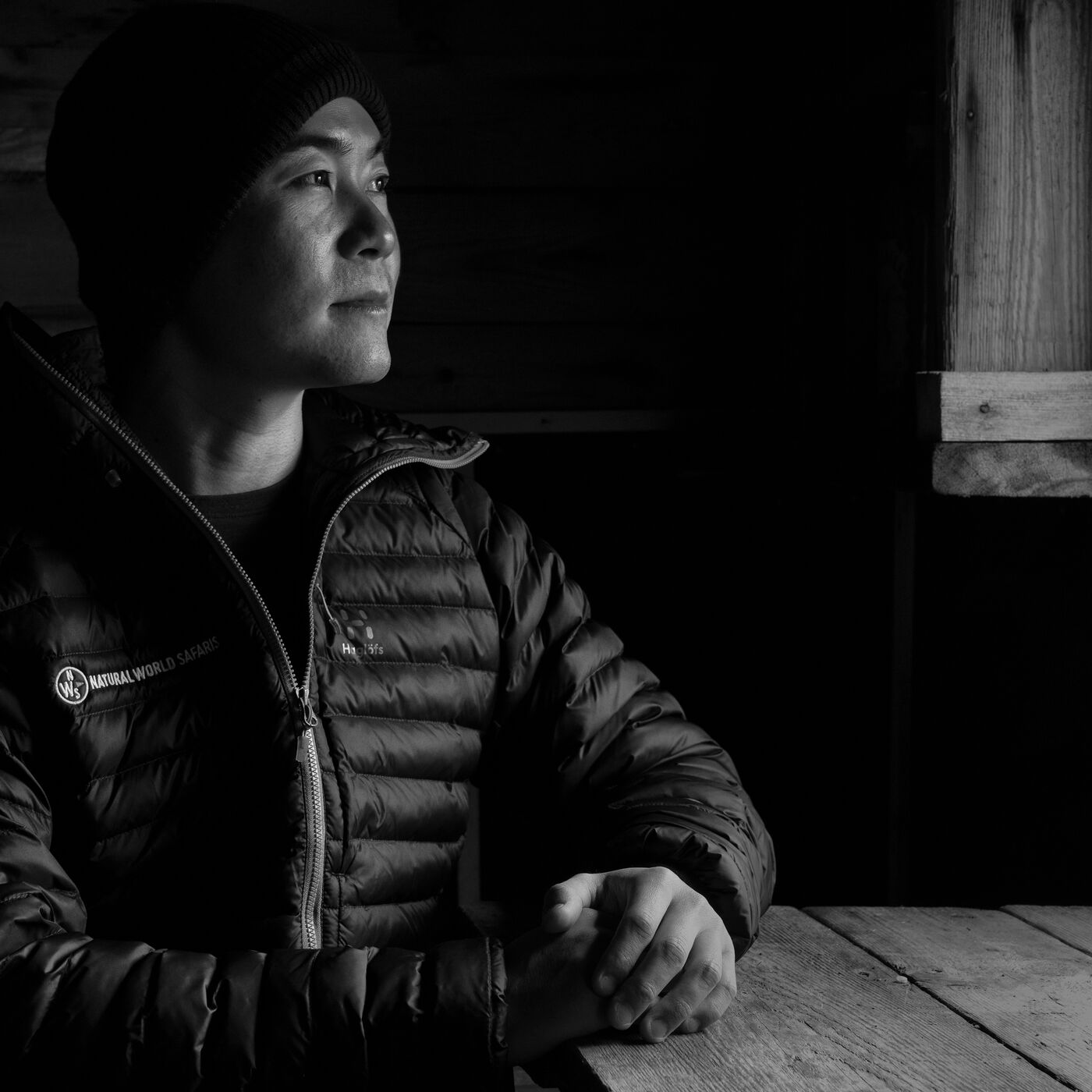Black-and-white portrait of a man in a beanie and down jacket inside the trapper's cabin, looking toward the window