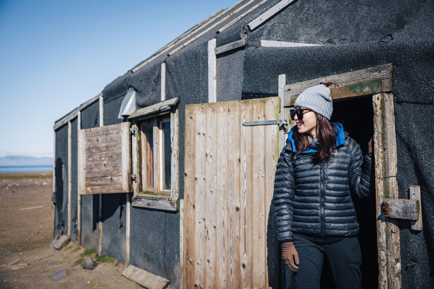 Grace in a beanie and black puffer leaning in the doorway of a weathered trapper's cabin at Andréetangen