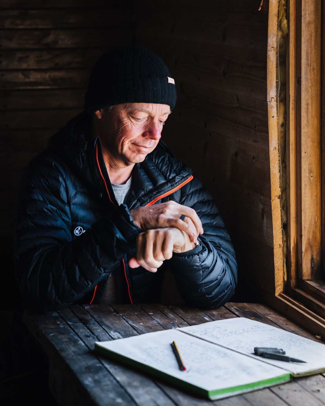 Man in a black beanie at the cabin table beside the logbook, adjusting his watch