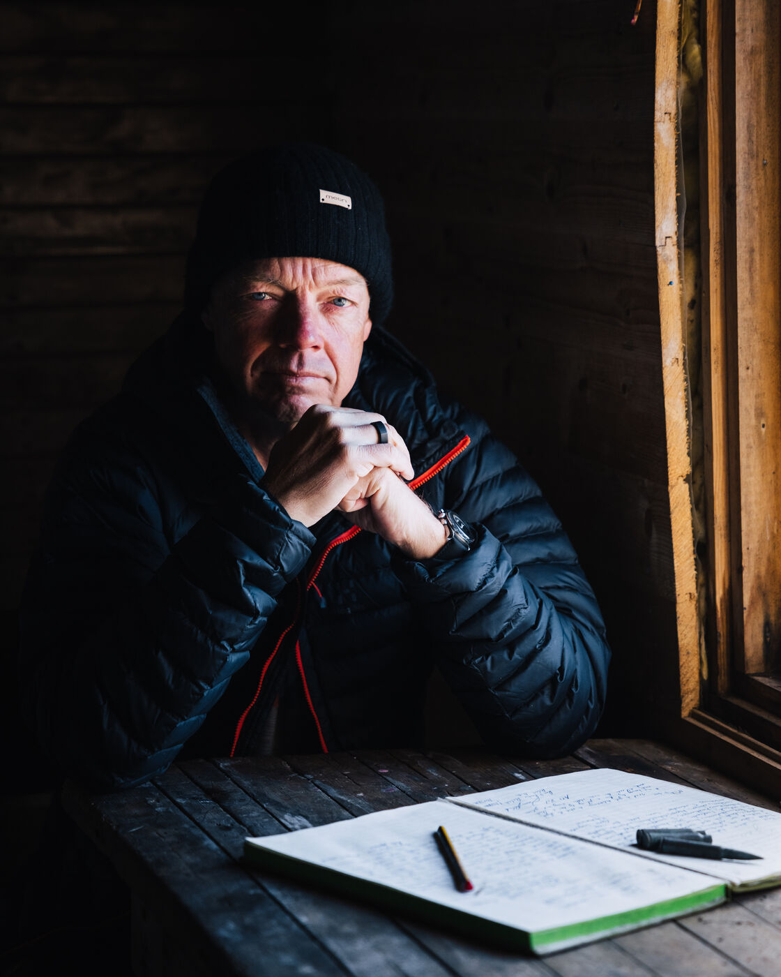 Man in a black beanie resting his chin on his hands at the cabin table, logbook in front