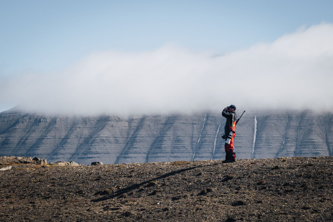 Guide with a rifle standing on the tundra below a cloud-capped, snow-striped mountain