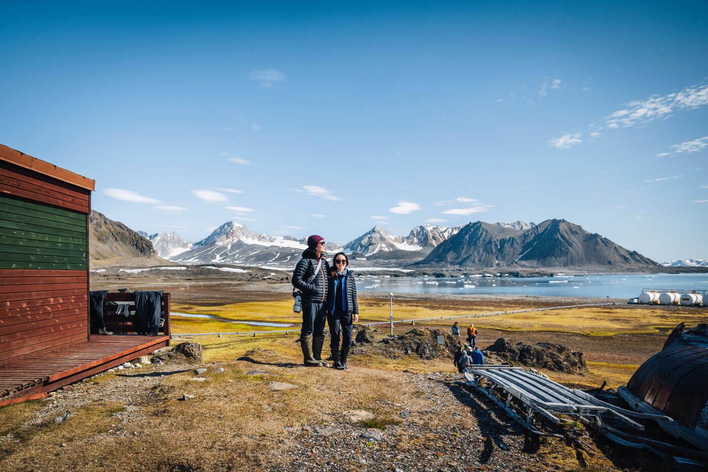 Two people standing outside a green station building, mountains and the fjord behind