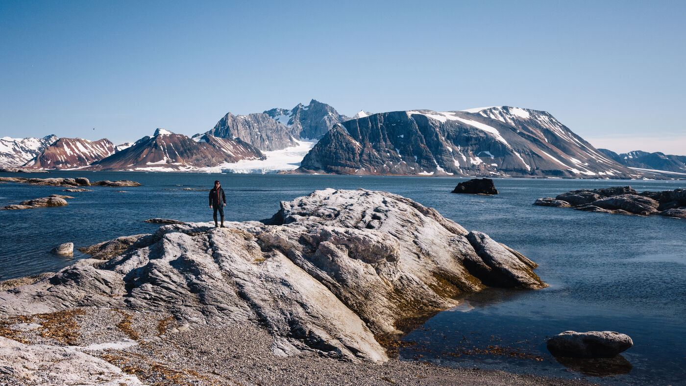 Lone figure standing on a rock outcrop at the water's edge, snow-capped mountains beyond
