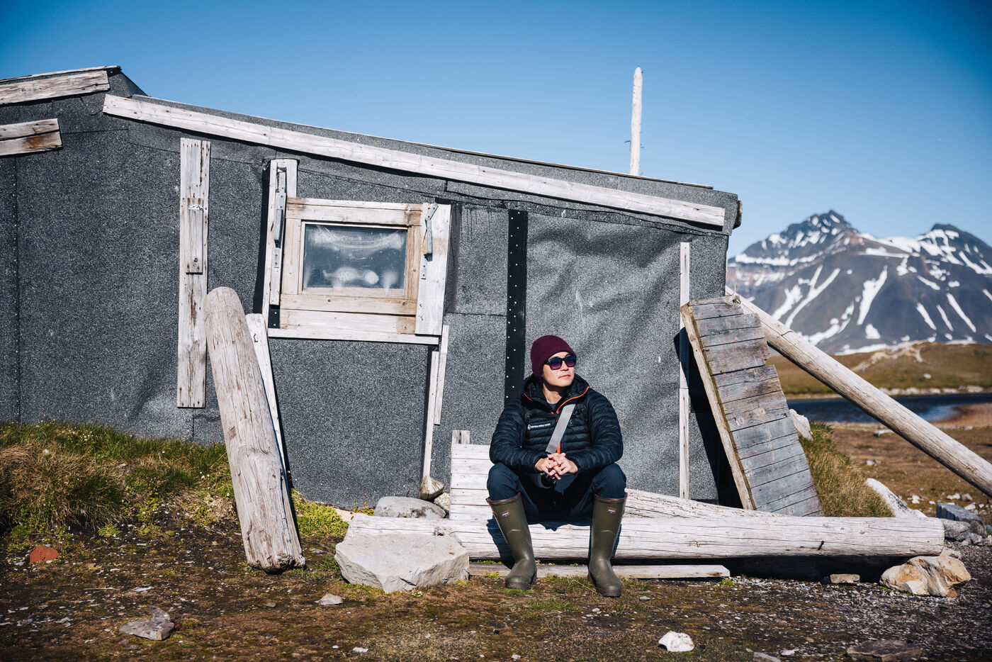 Woman in a maroon beanie sitting on driftwood outside a weathered tar-paper cabin, mountains behind