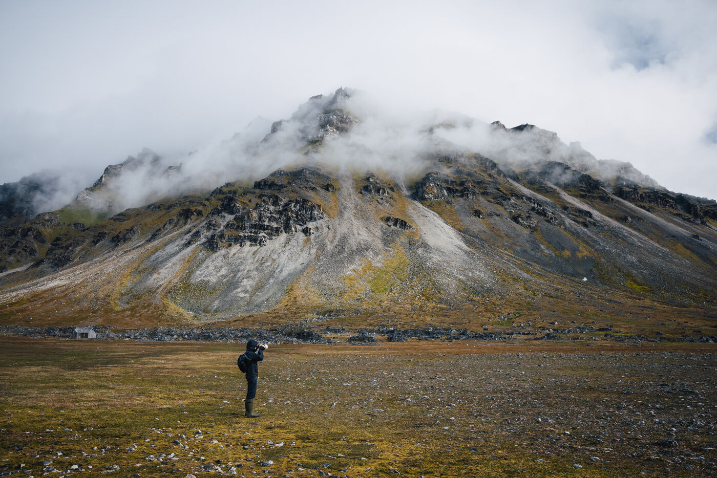 Lone figure standing on the tundra below a cloud-shrouded mountain