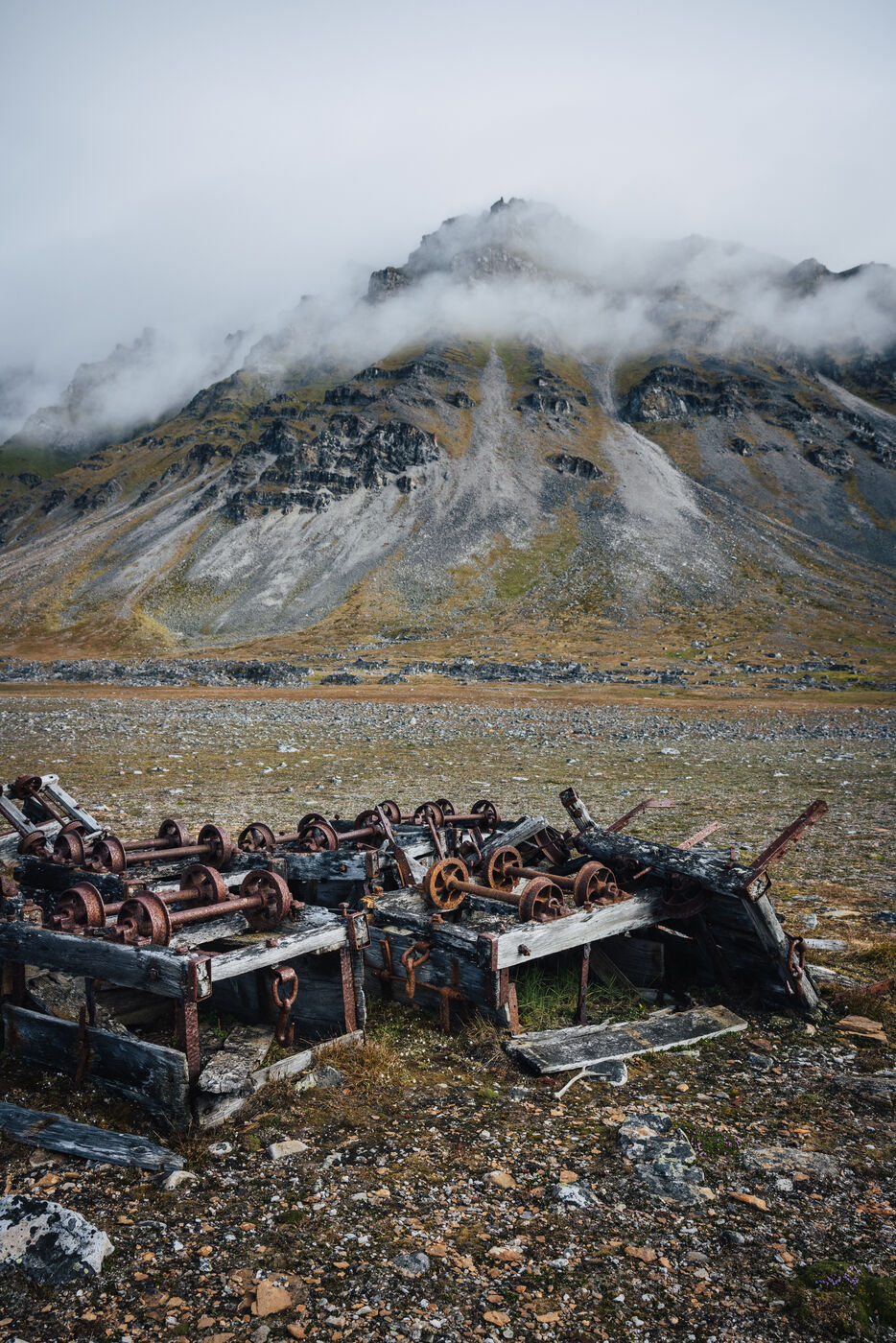 Rusted wheels and weathered timber of abandoned machinery on the tundra below a cloud-capped mountain