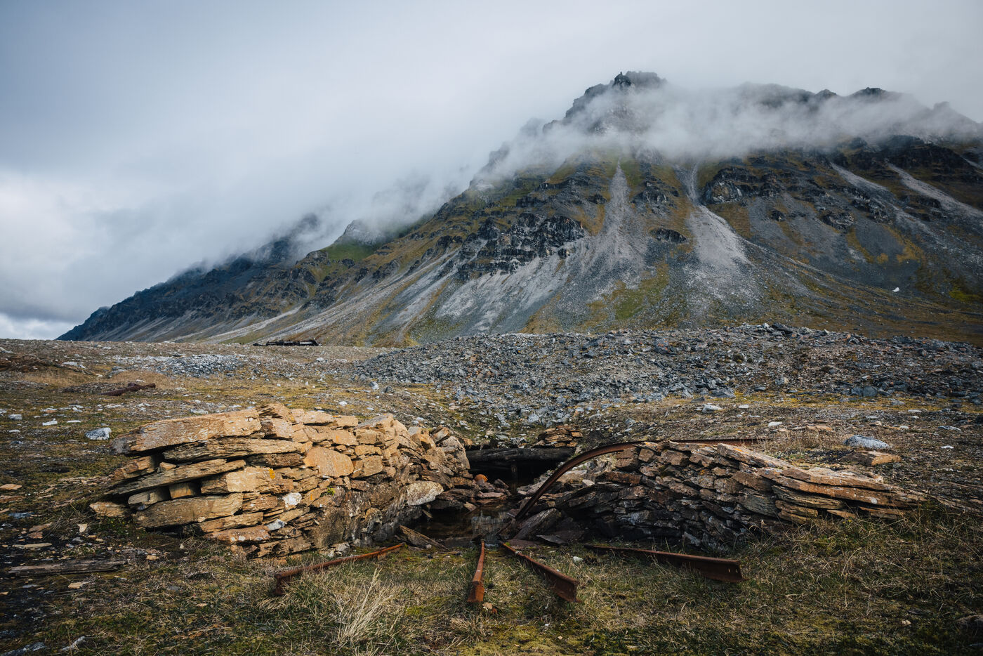 Collapsed stone structure with rusted ironwork on the tundra below a cloud-capped mountain