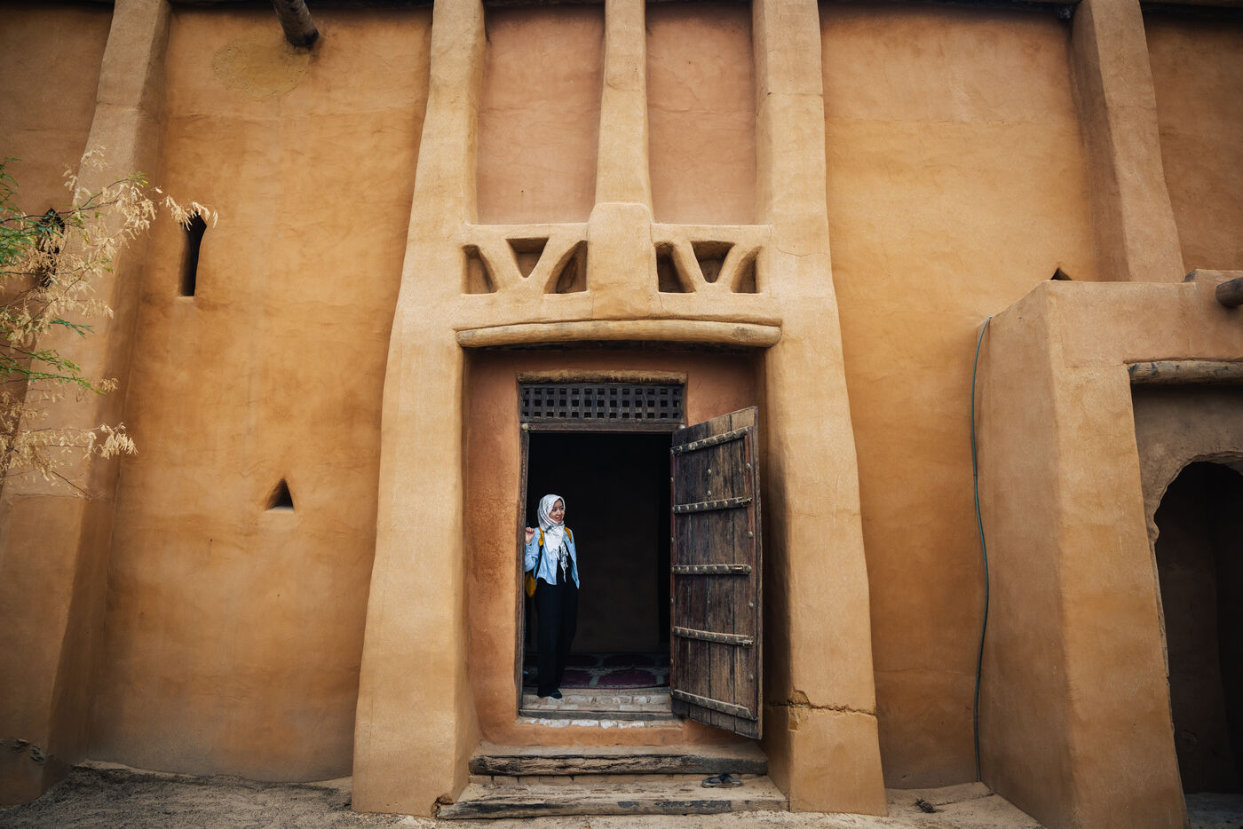 Person standing in an ornate mud-plastered mosque doorway with a carved wooden door open beside her
