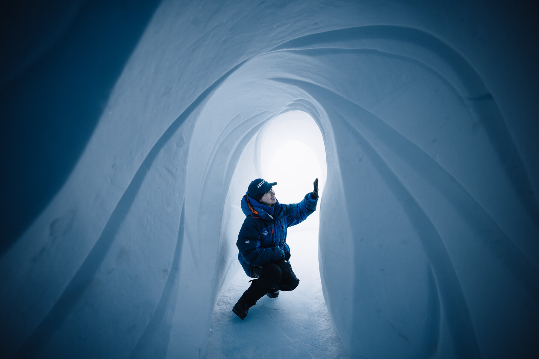 Man in a blue parka inside a curved ice tunnel at Wolf's Fang reaching up toward the glowing blue ceiling