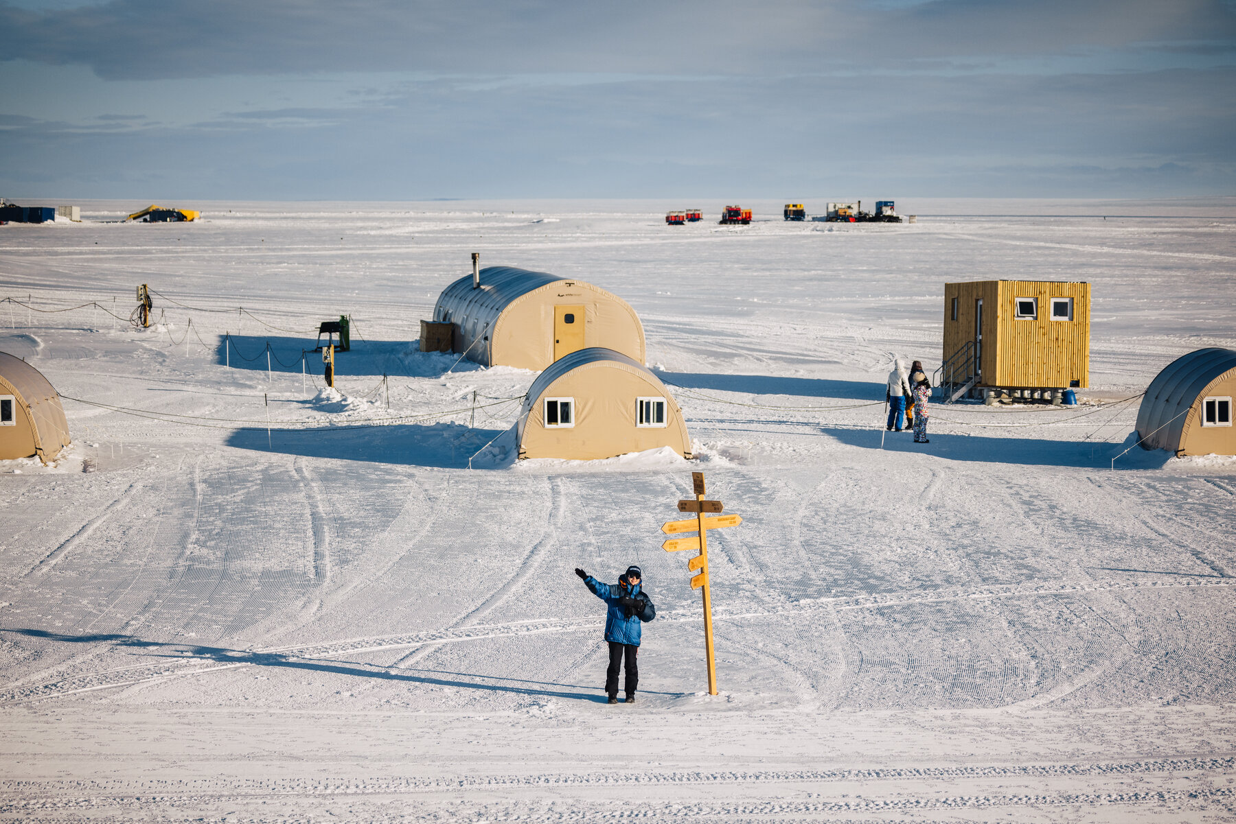 Wolf's Fang camp on the Antarctic plateau with yellow Quonset tents, a wooden directional signpost, and a figure in a blue parka on the endless white horizon