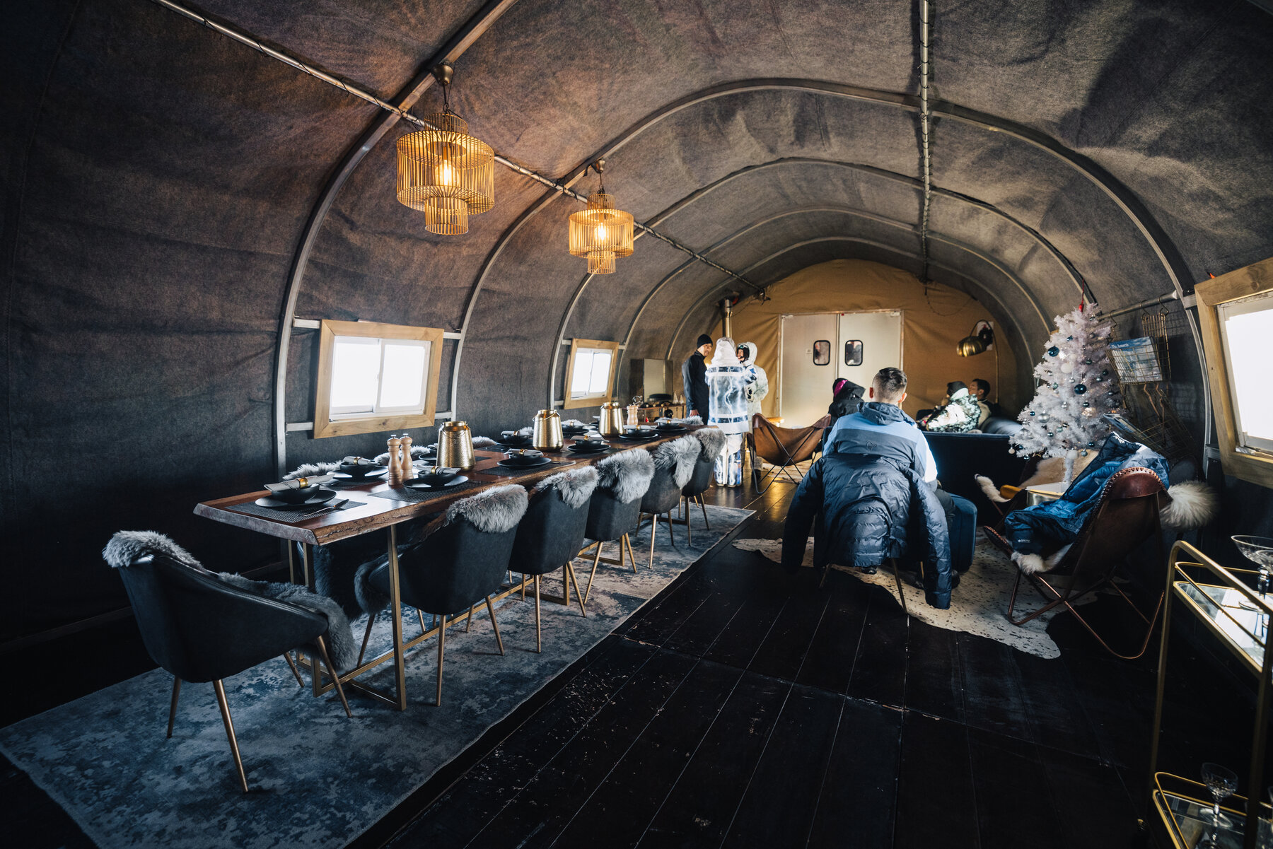 Interior of Wolf's Fang main lodge with a long wooden dining table, sheepskin-draped chairs, pendant lighting, and dark canvas arched walls