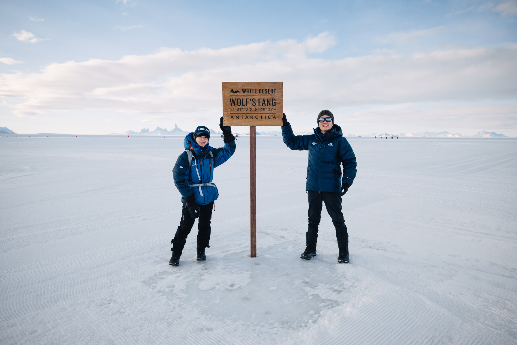 Jason and Grace standing beside the wooden Wolf's Fang White Desert Antarctica signboard on the plateau