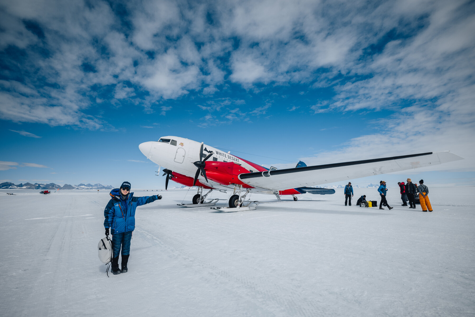 Ski-equipped Basler BT-67 in White Desert livery parked on the snow with a mountain range on the horizon and a passenger in a blue parka pointing toward it