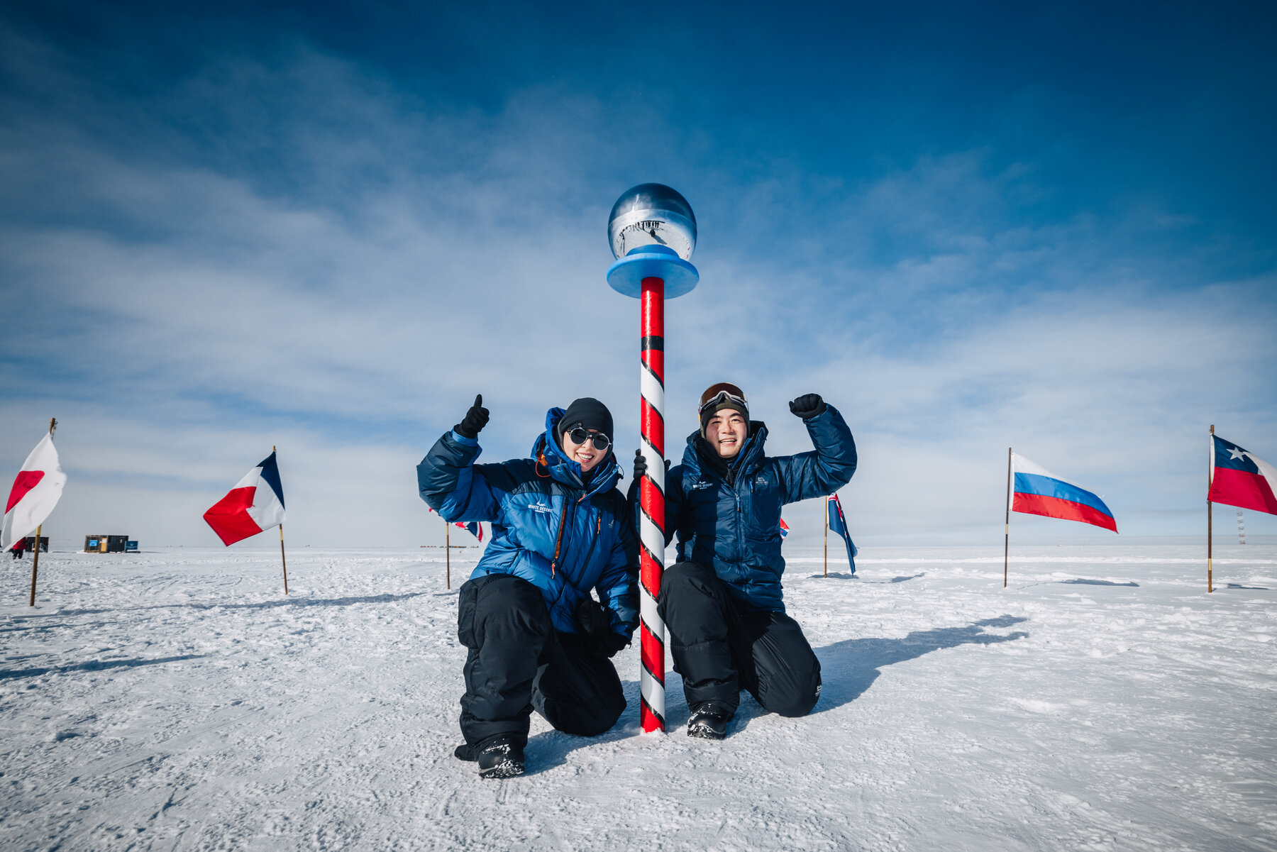 Jason and Grace kneeling together at the ceremonial South Pole marker with international flags behind them