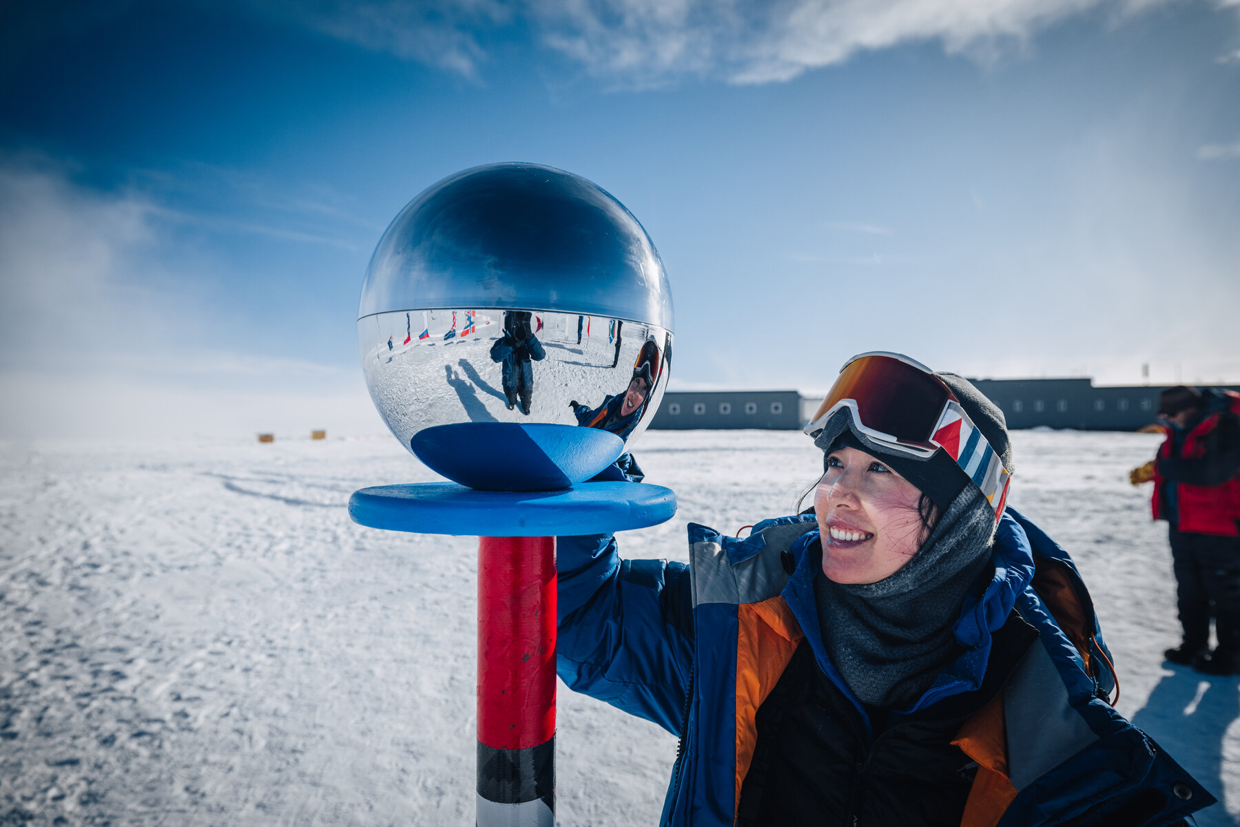 Grace smiling up at the mirrored silver sphere of the ceremonial South Pole marker with the elevated station in the background