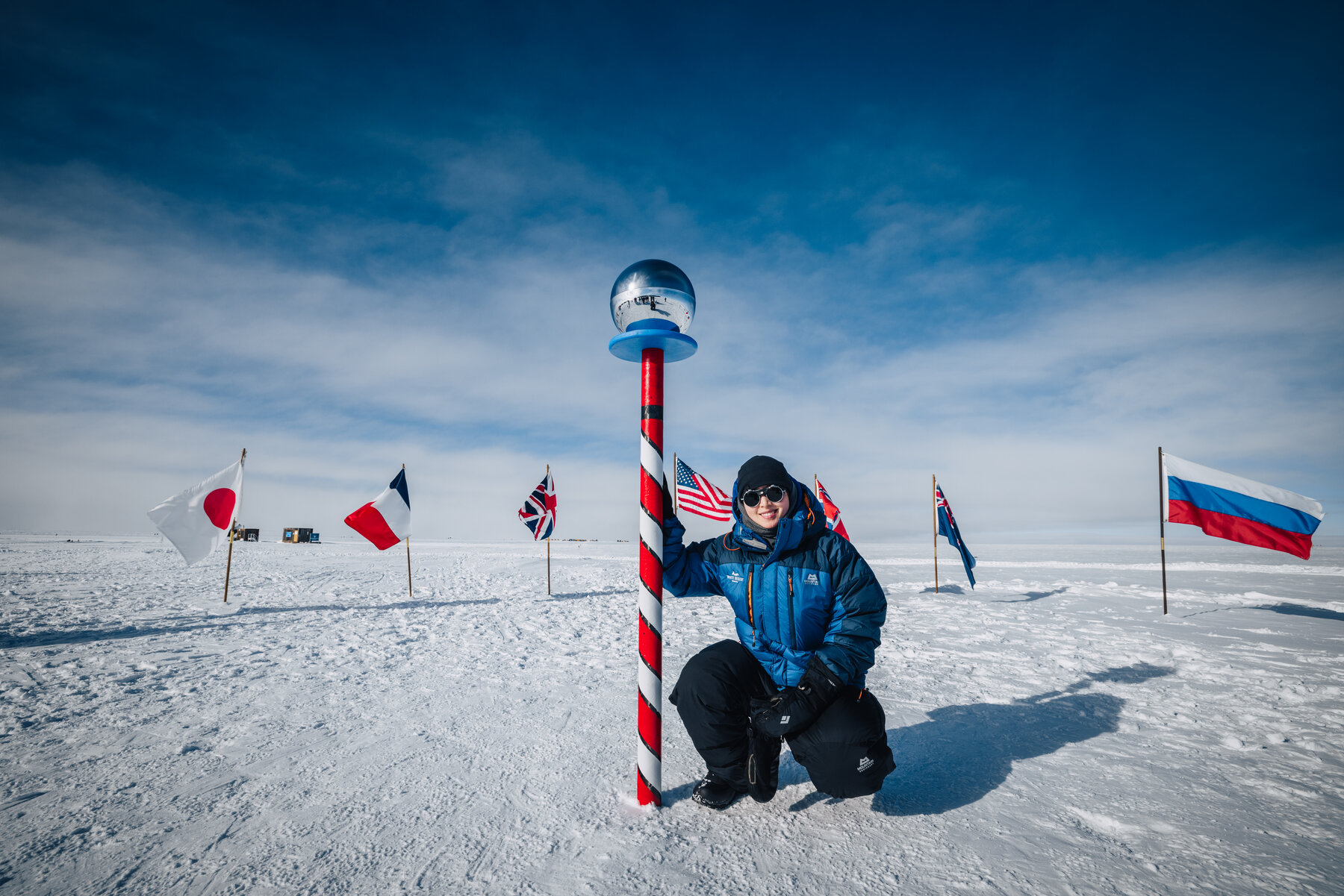 Jason standing alone at the ceremonial South Pole marker with the flags of the Antarctic Treaty nations behind him