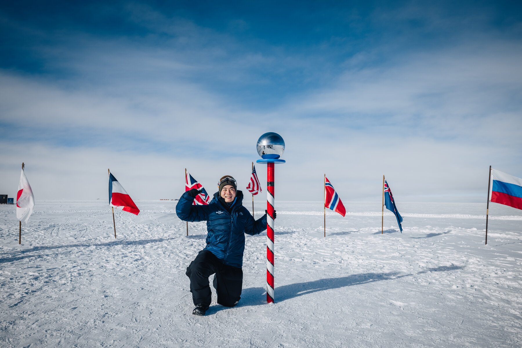 Jason kneeling beside the ceremonial South Pole marker with its polished silver sphere surrounded by the flags of Antarctic Treaty nations