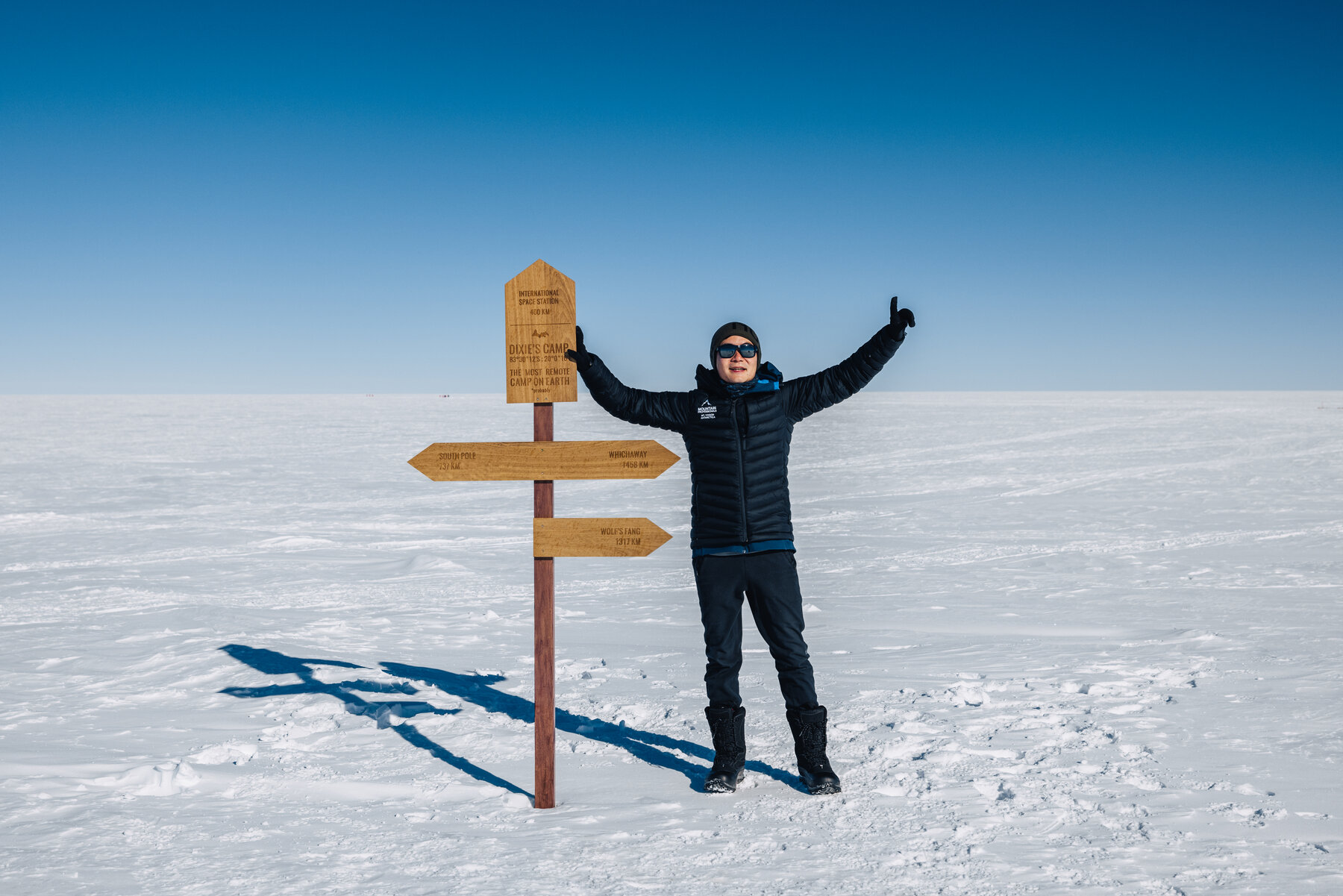 Jason with arms raised at the wooden Dixie's Camp directional signpost on the polar plateau