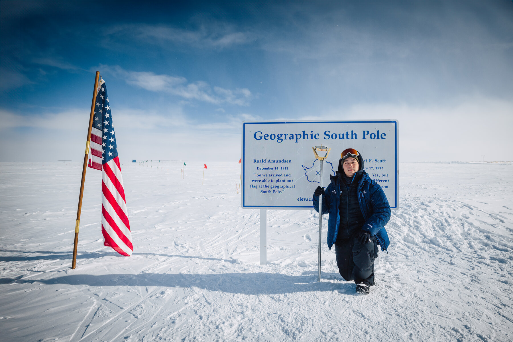 Jason in a navy parka standing beside the Geographic South Pole sign at 90 degrees south with the American flag and ice plateau behind