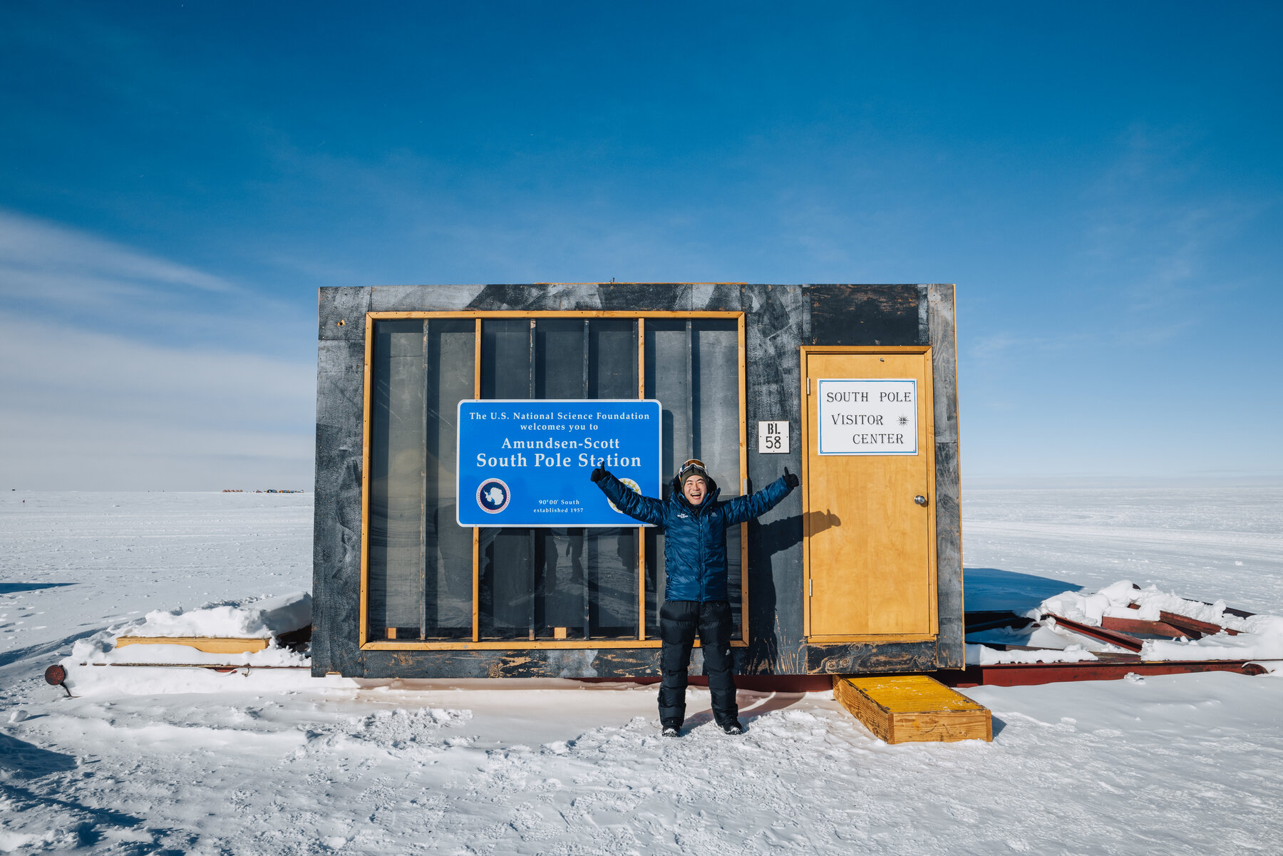 Jason posing with arms out in front of the small South Pole Visitor Center hut with its blue Amundsen-Scott welcome sign and yellow door