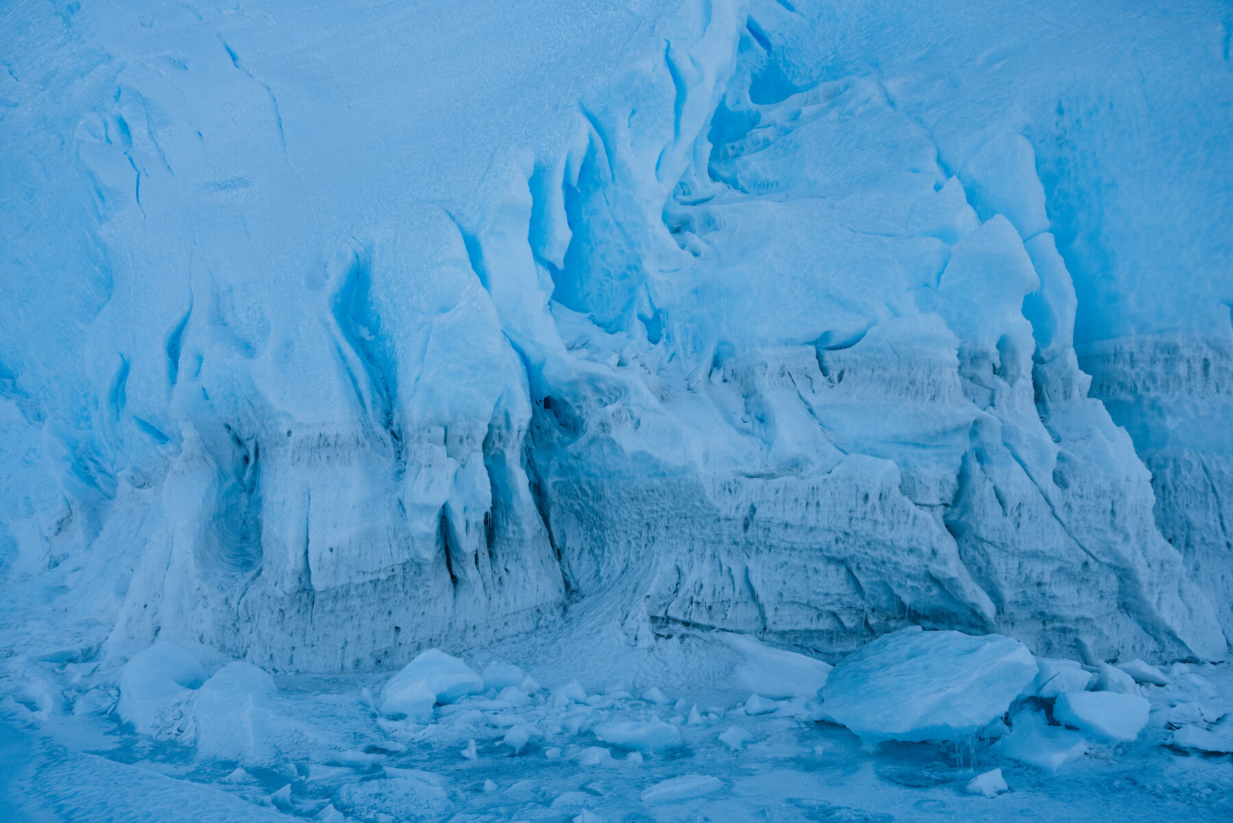 Close-up detail of the carved blue glacial ice cliff face with deep fissures, ledges, and iceberg fragments at the base