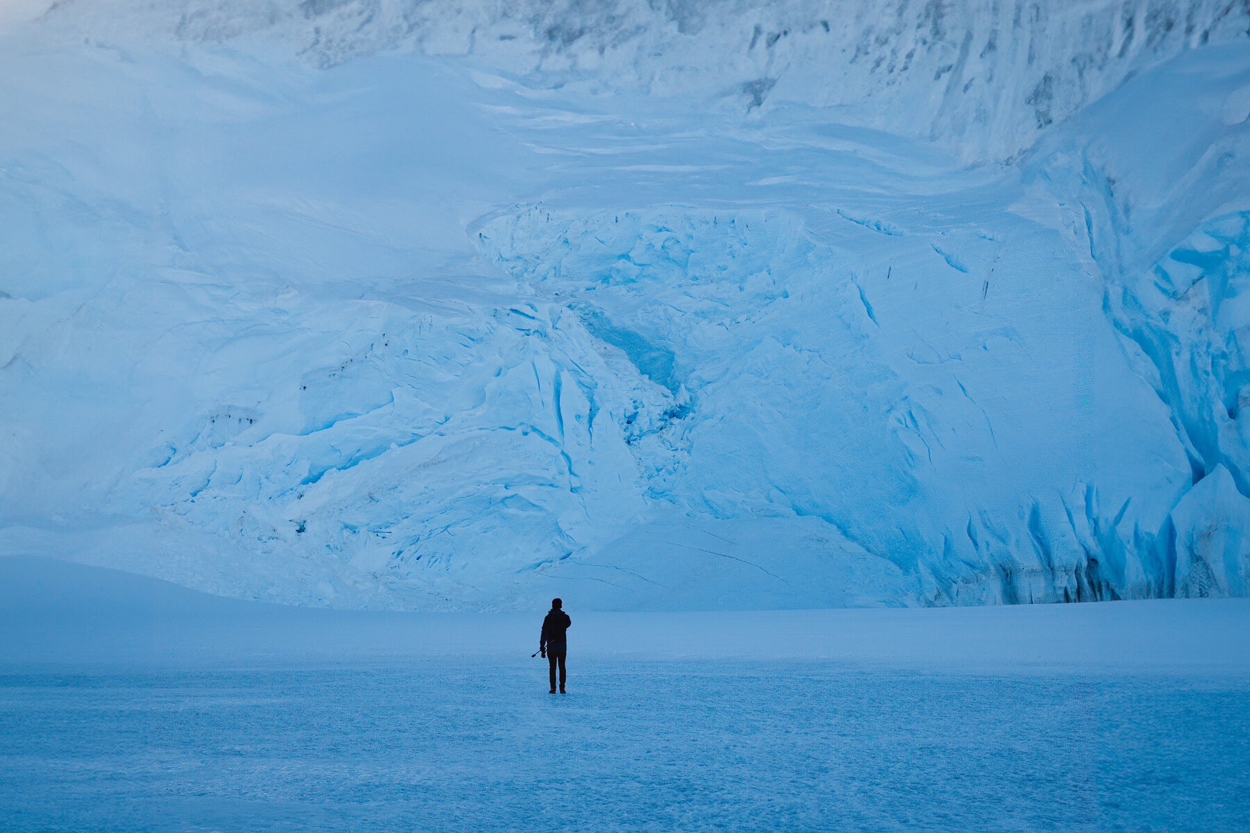 Solitary figure standing on a snowfield at the base of a towering blue ice cliff, dwarfed by the wall of glacier behind