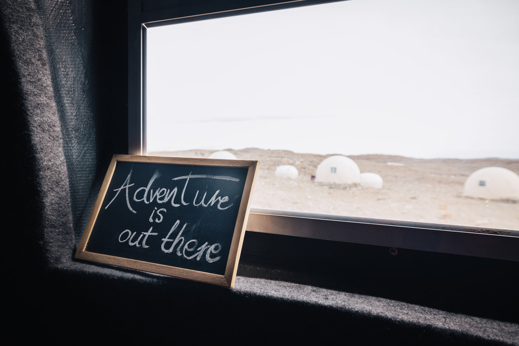 Small framed chalkboard reading 'Adventure is out there' resting on a windowsill looking out to the Whichaway dome pods and ice plateau