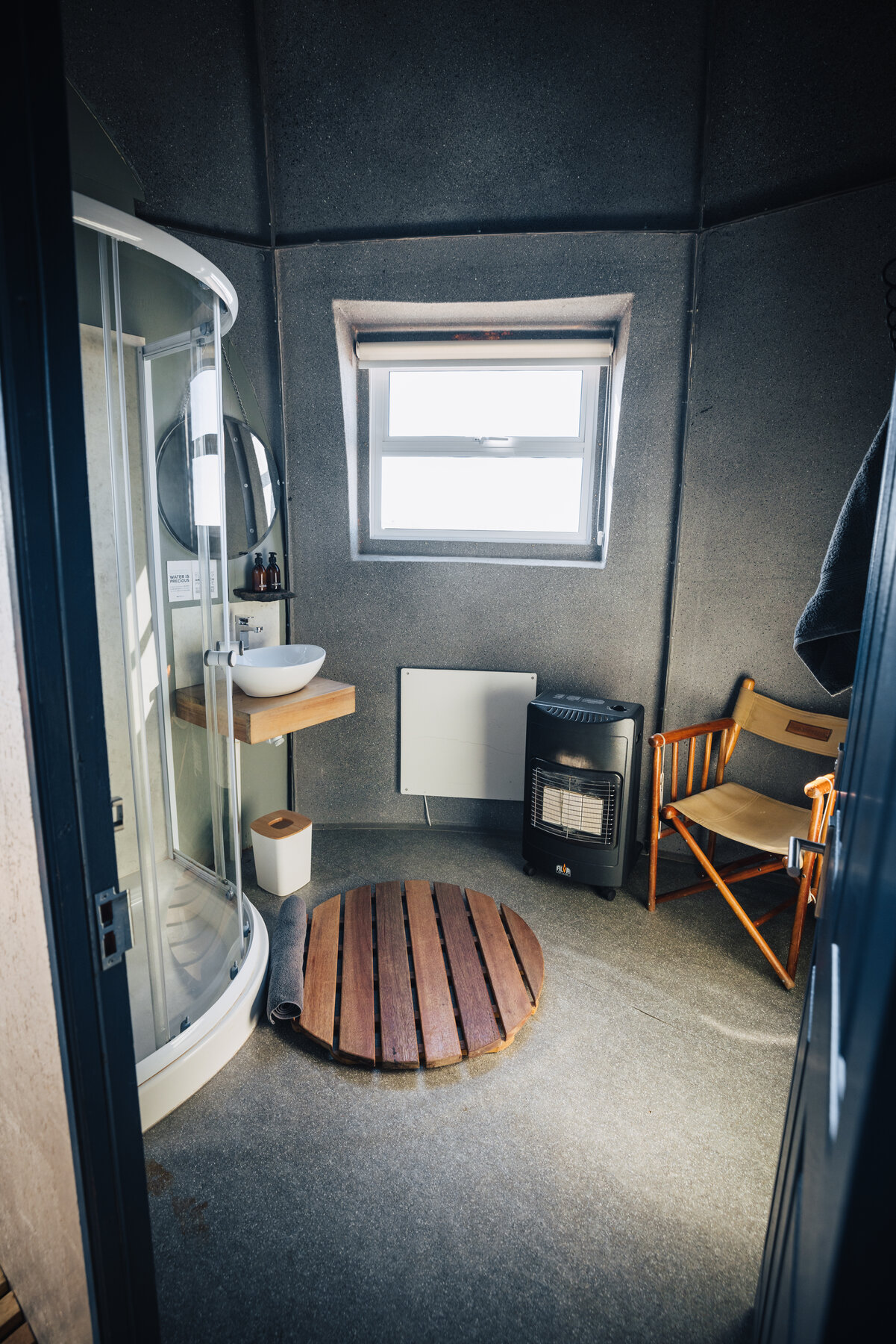 Whichaway pod bathroom with corner shower cabinet, circular teak bathmat, wall heater, and folding wooden chair