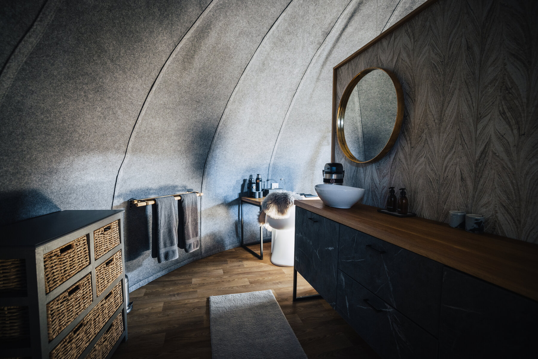 Modern pod bathroom with a curved felt wall, round brass-framed mirror, wooden countertop vanity, vessel sink, and woven storage baskets underneath