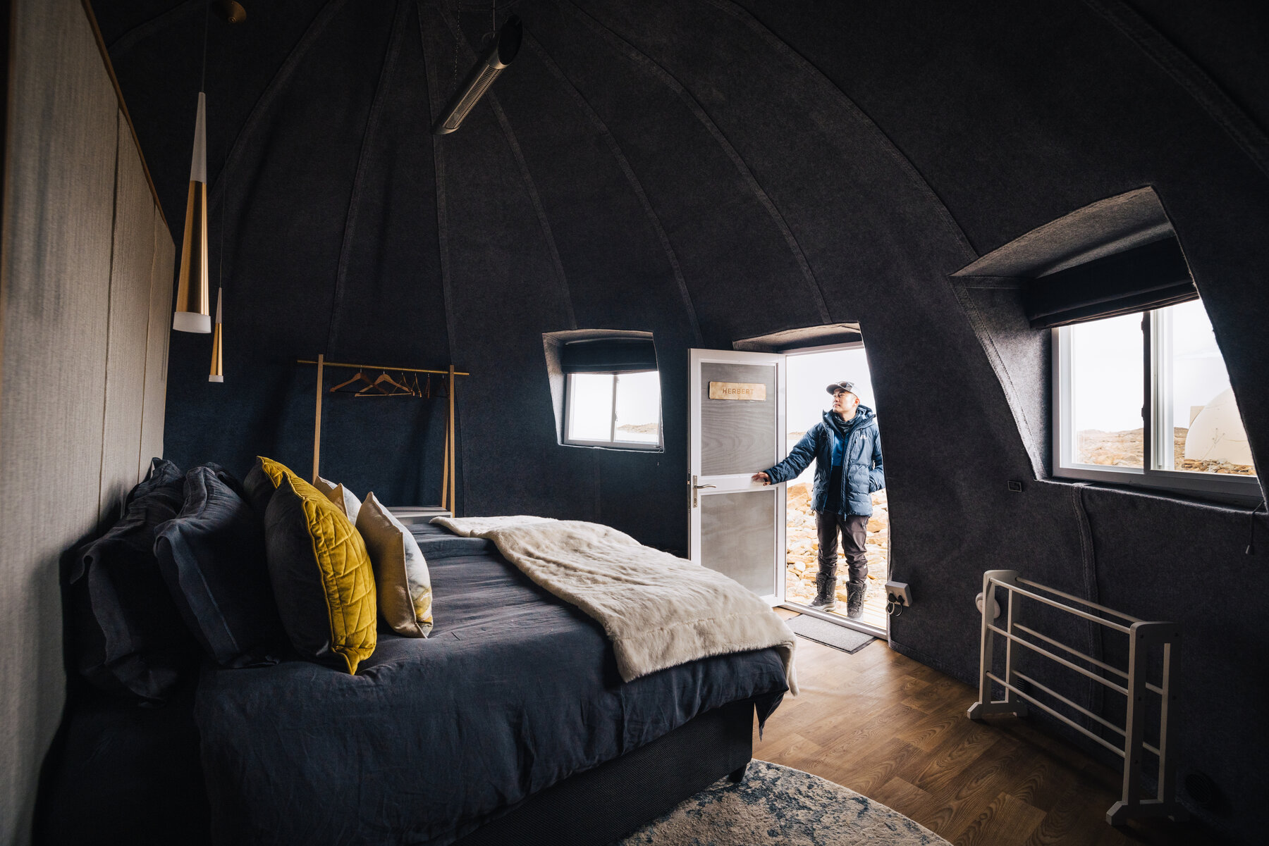 Jason standing in the open doorway of a Whichaway sleeping pod backlit by daylight, with a dark-domed ceiling, a large bed with yellow pillows, and polished wood floors