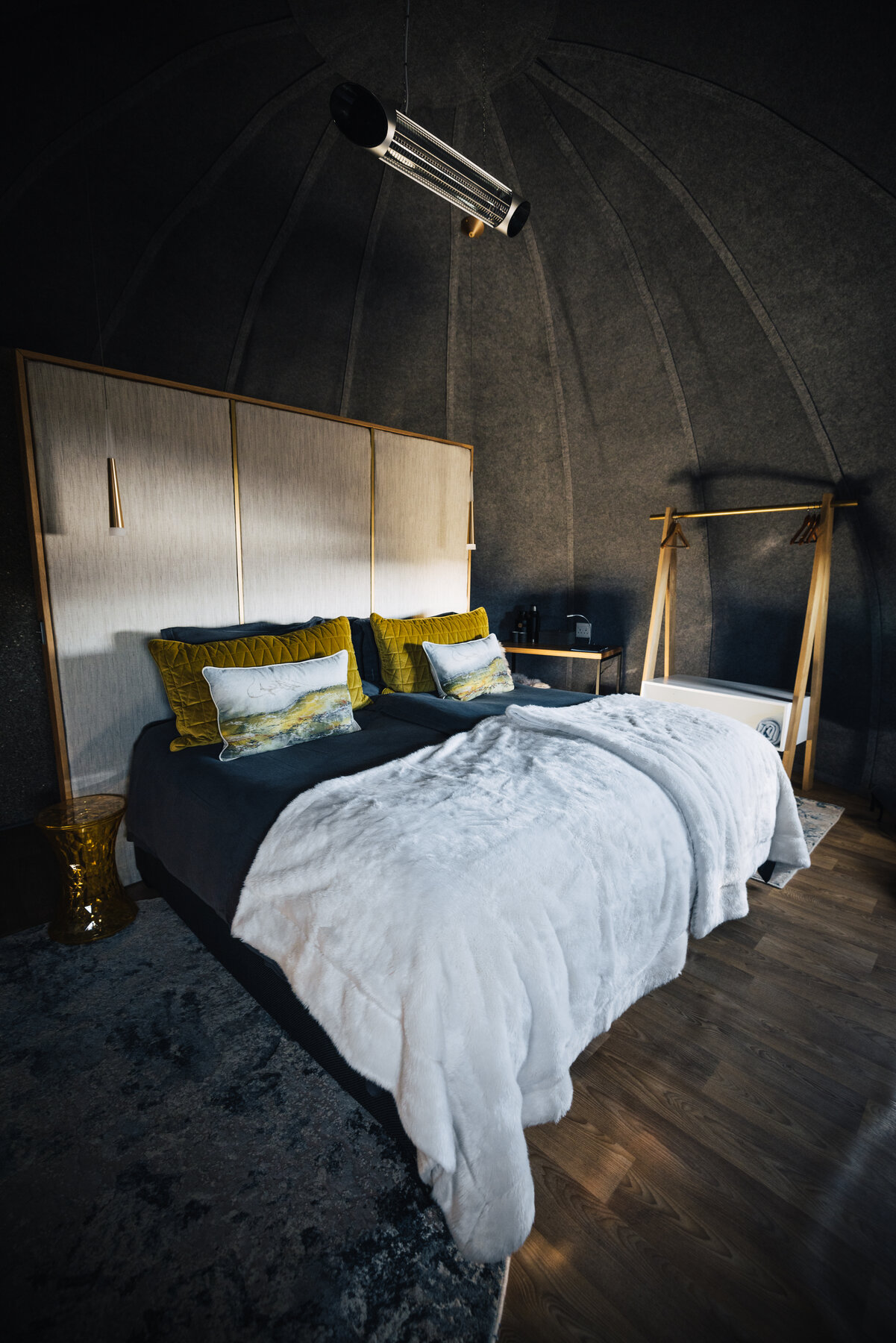 Portrait view of a Whichaway pod bedroom with dramatic low lighting, brass chrome pendant lamp, and a plush bed with yellow pillows