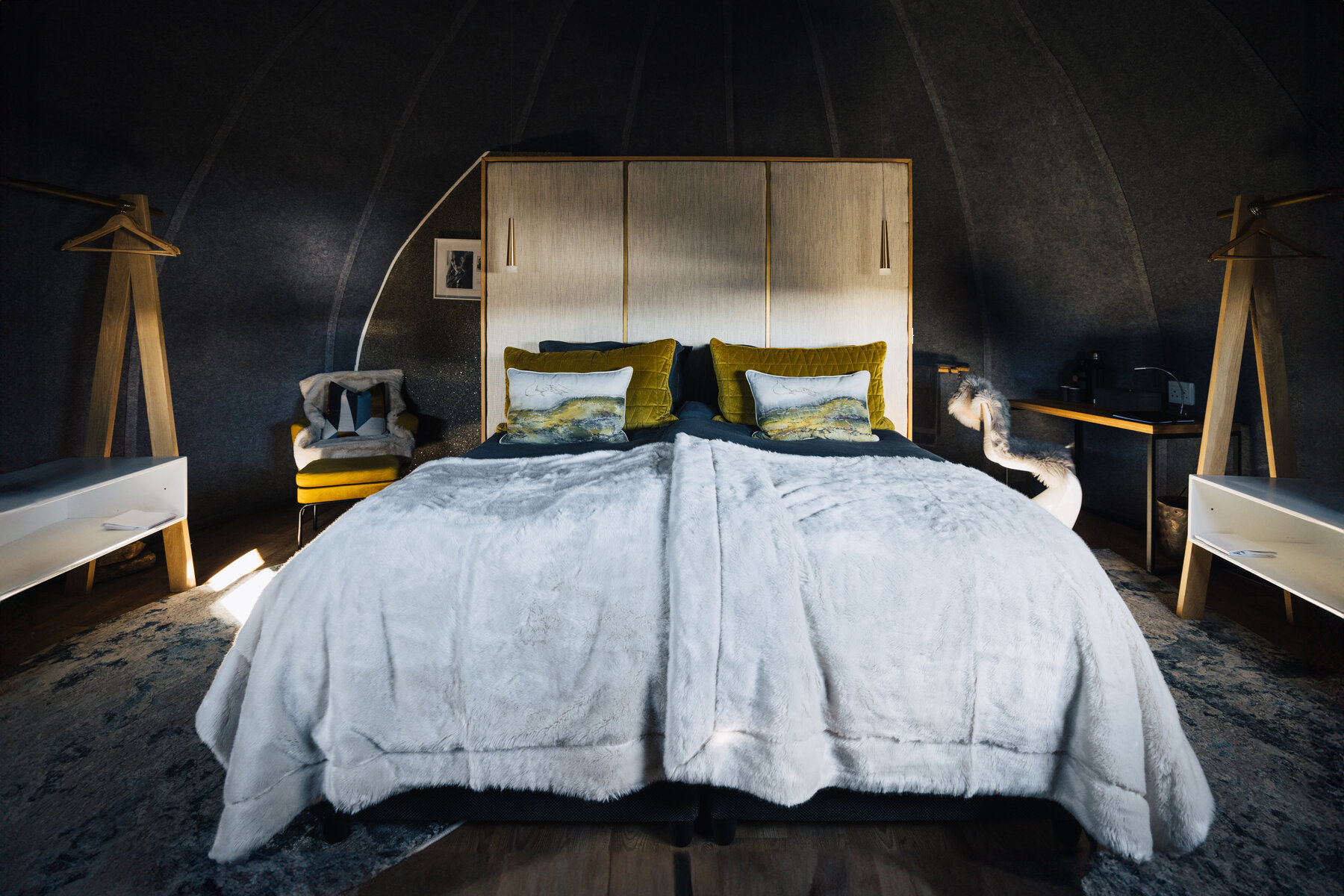 Wide view of a Whichaway bedroom pod with a king bed dressed in dark linens and yellow velvet pillows, a wooden headboard, and backlit side tables with reading lamps