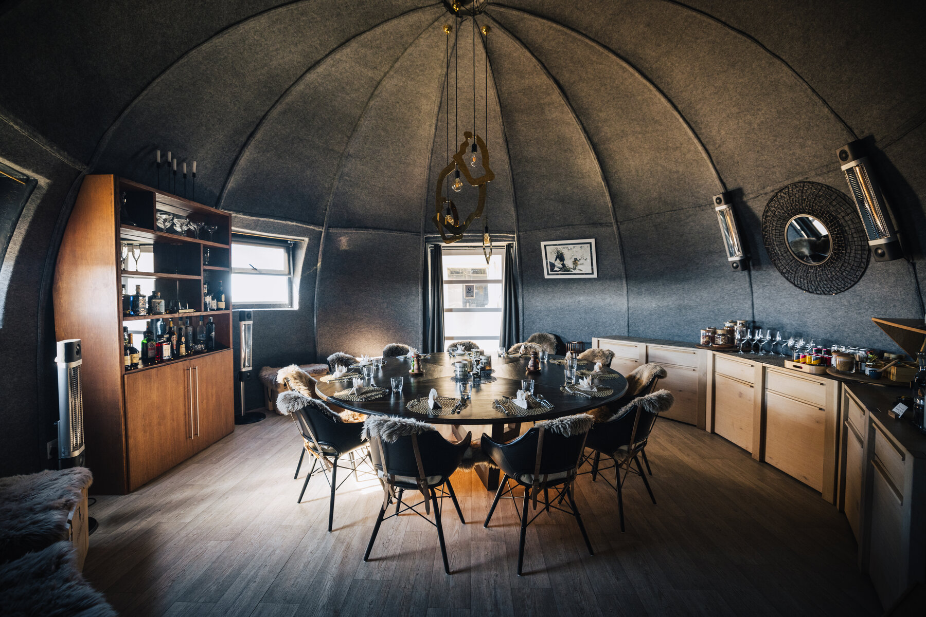 The Whichaway dining pod with a round wooden table surrounded by sheepskin-draped chairs, pendant lighting, a wine bar, and dark felt-lined dome walls