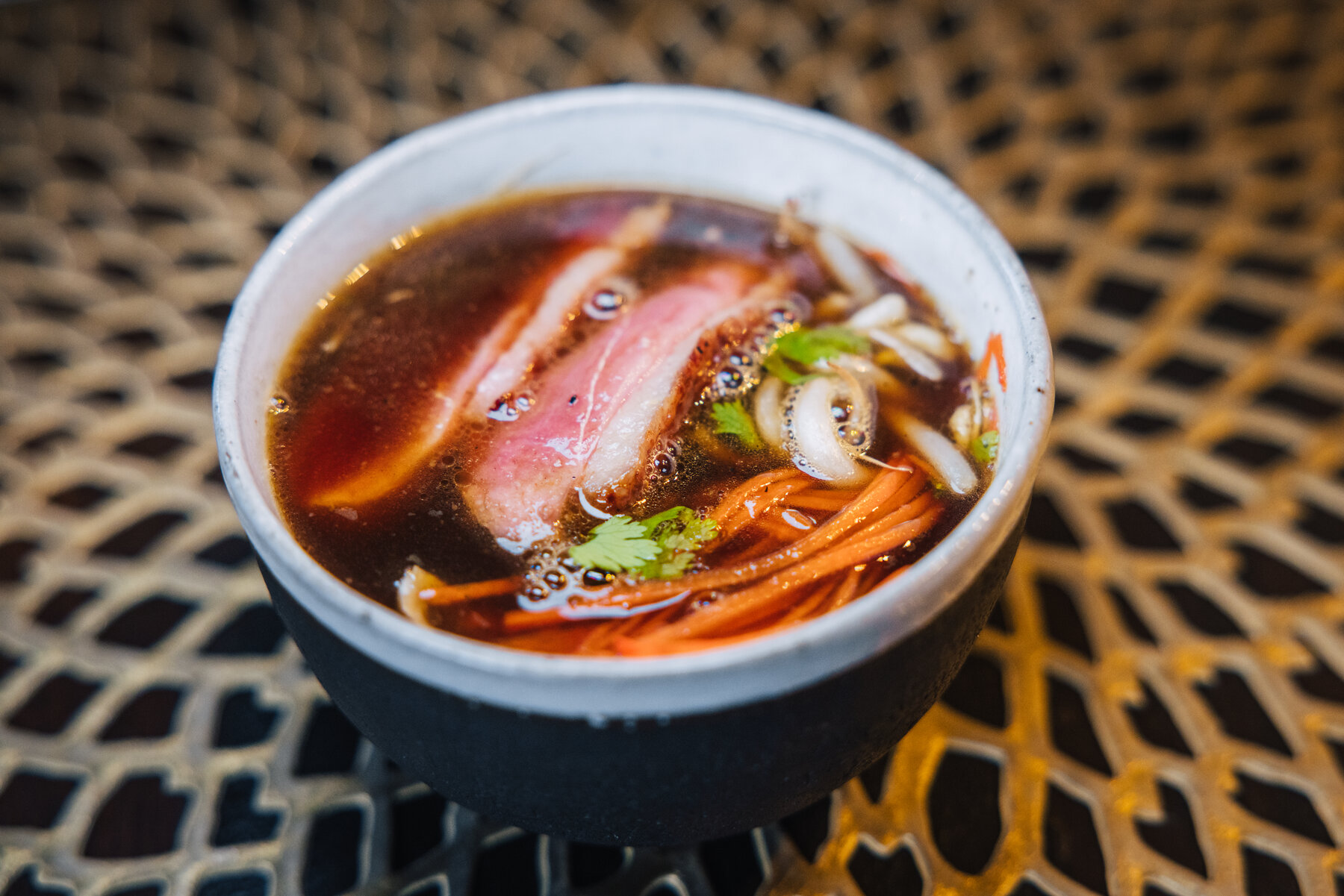 A small black bowl of clear consommé with seared duck breast, carrot threads, and cilantro at the Whichaway dining pod